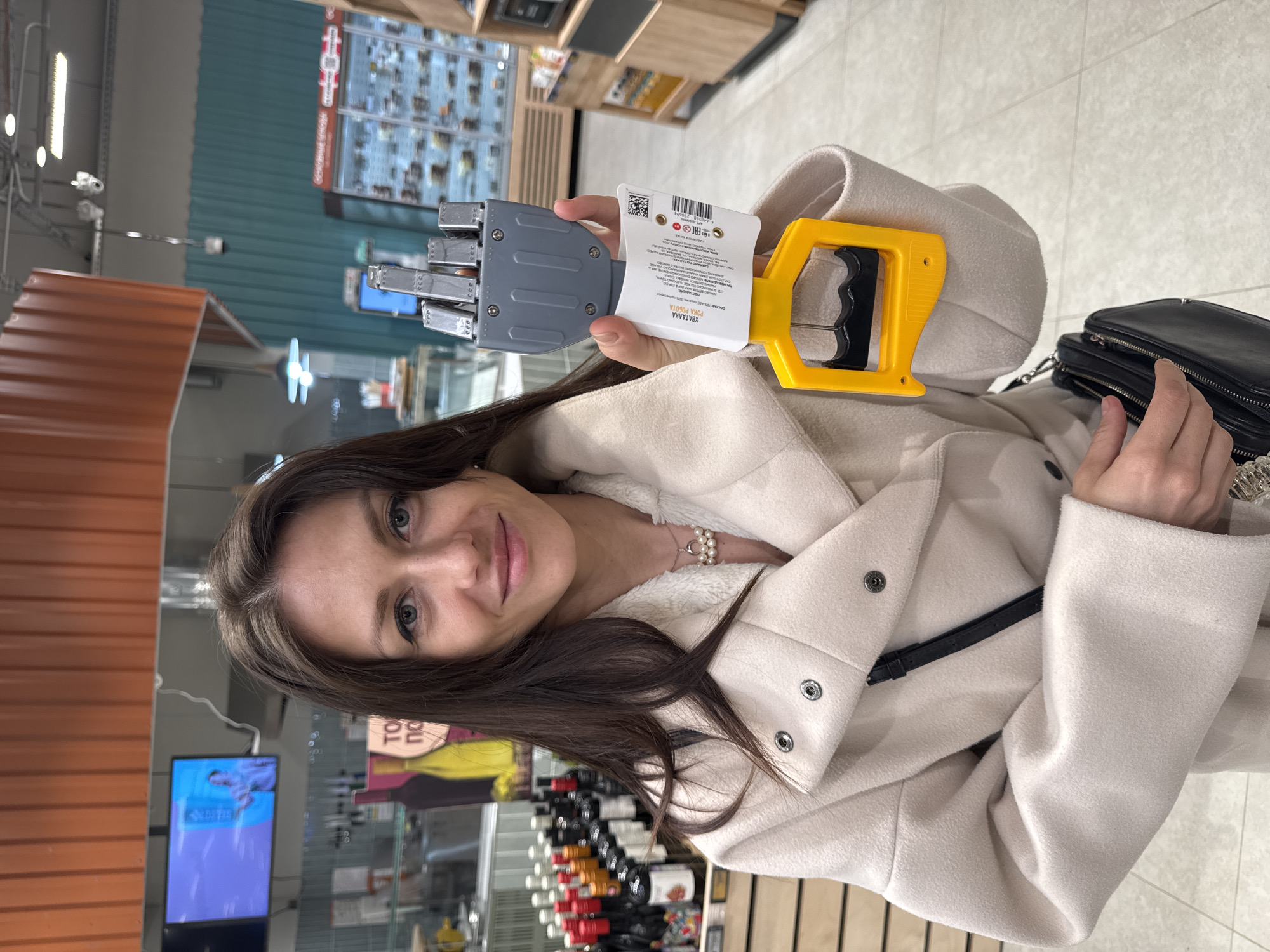 A woman in a beige coat and a black purse is holding a yellow and gray grabbing tool, likely a grabber, in a store. Rows of wine bottles are visible behind her. The setting appears to be a grocery store or a store that sells food items.