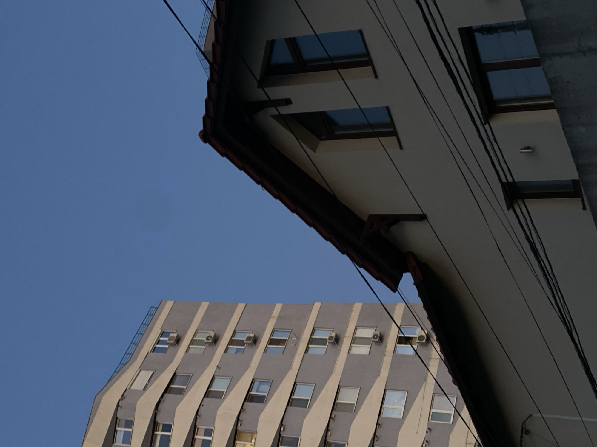 The image depicts an urban scene featuring two buildings against a clear blue sky. On the left, a tall, multi-story apartment building with a grey and beige striped facade is visible. Several windows with white frames and air conditioning units are sparsely distributed across its height. To the right and closer to the foreground, a smaller, cream-colored building with a red tile roof is partially visible. This building has dark-framed windows, including a dormer window. Dark electrical wires crisscross the scene in front of both structures. The setting is likely Chișinău, Moldova, given the architectural style and the general urban context. There are no people or animals visible in the image.
