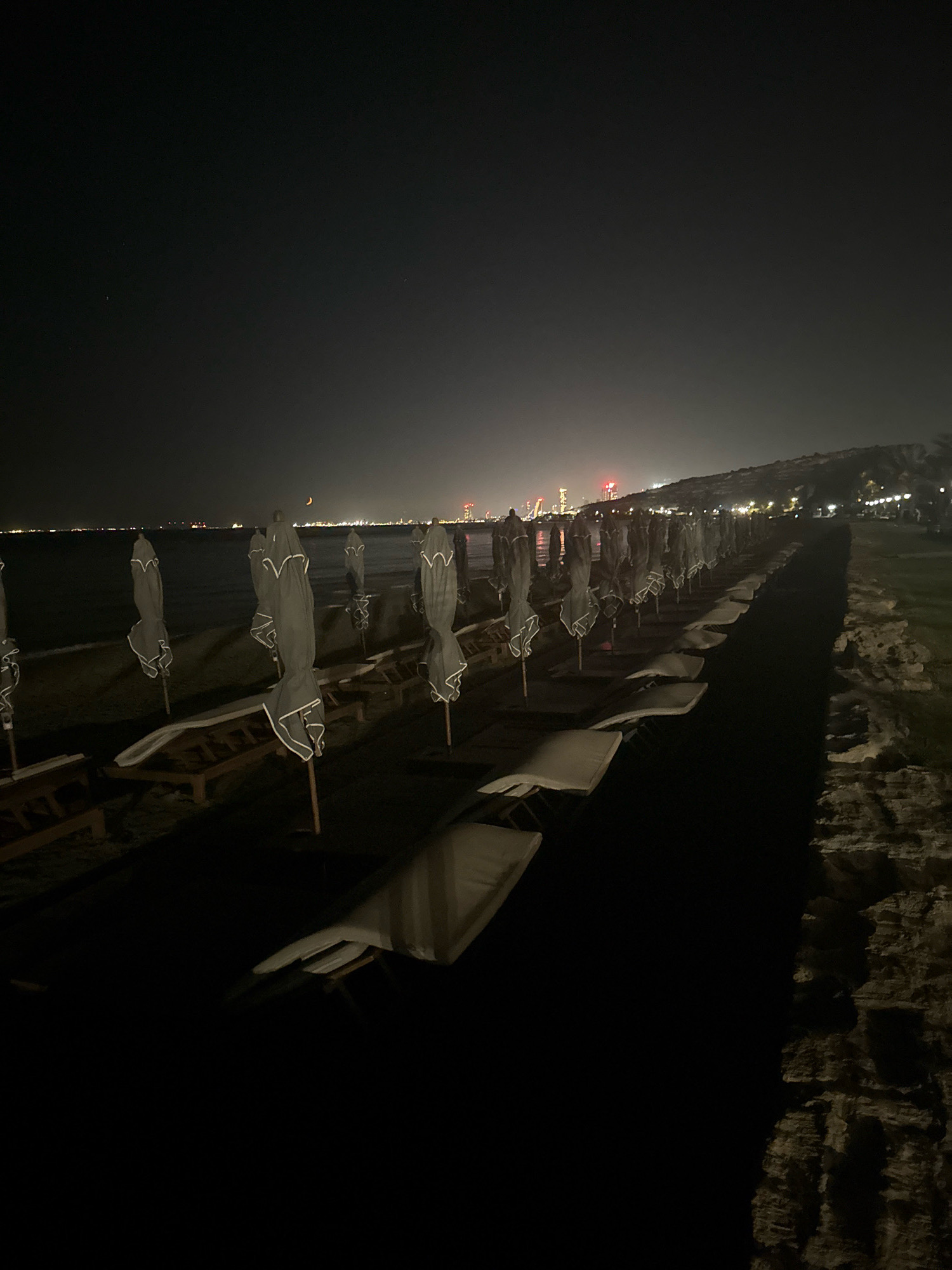An outdoor, nighttime, wide-angle shot of a beach in Agios Tychona Community, Cyprus, features a long row of closed beach umbrellas and lounge chairs lined up on the sand. The dark sea stretches out to the horizon, where a distant city skyline glows with lights. A faint crescent moon is visible in the dark sky above. The overall atmosphere is quiet and serene, with the gentle lapping of waves against the shore.