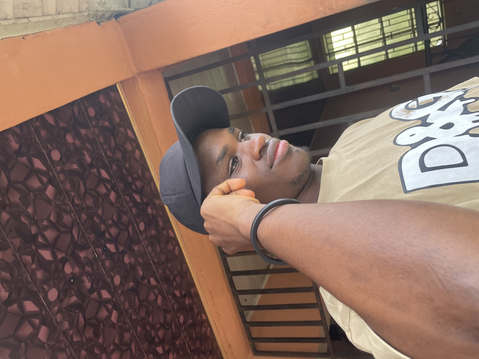 A young Black male is pictured from the chest up, outdoors, wearing a tan t-shirt with the letters "D&G" in white, and a black baseball cap. He is looking to his right, with his right hand touching his temple and forehead. A black wristband is visible on his left wrist. The background includes an orange-painted wall, a barred window with green reflections, and a patterned ceiling. The image is likely taken in Ihiagwa, Nigeria, given the architecture and common outdoor scenes depicted.