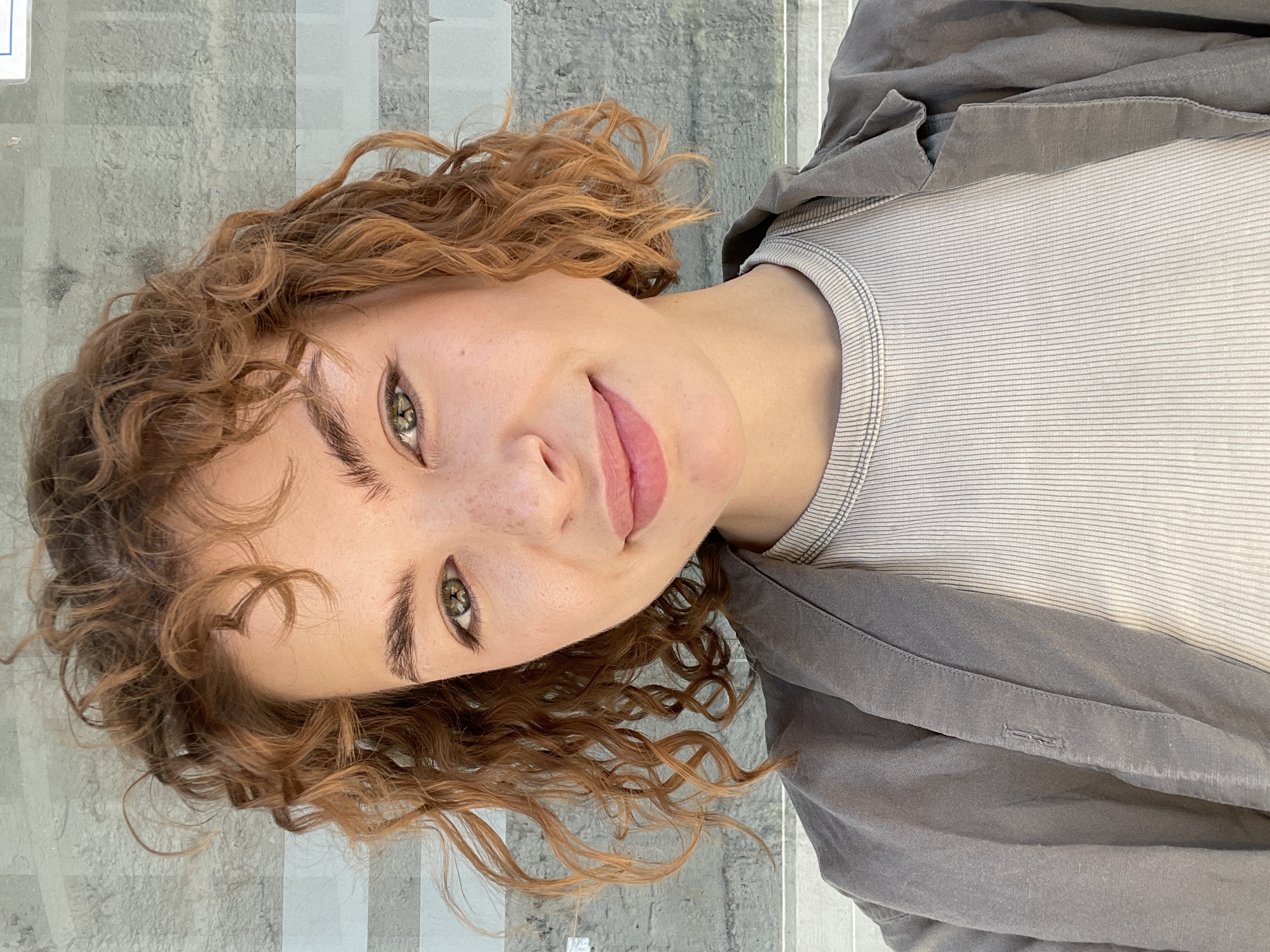 A young woman with curly red hair is seen from the chest up in a tilted, close-up portrait. She has fair skin with freckles and green eyes. She is wearing a light-colored, striped top and a gray jacket. The background appears to be a textured, gray surface with subtle white lines, possibly a stone or concrete wall with a glass or plastic barrier. The location is unknown and cannot be determined from the details within this image.
