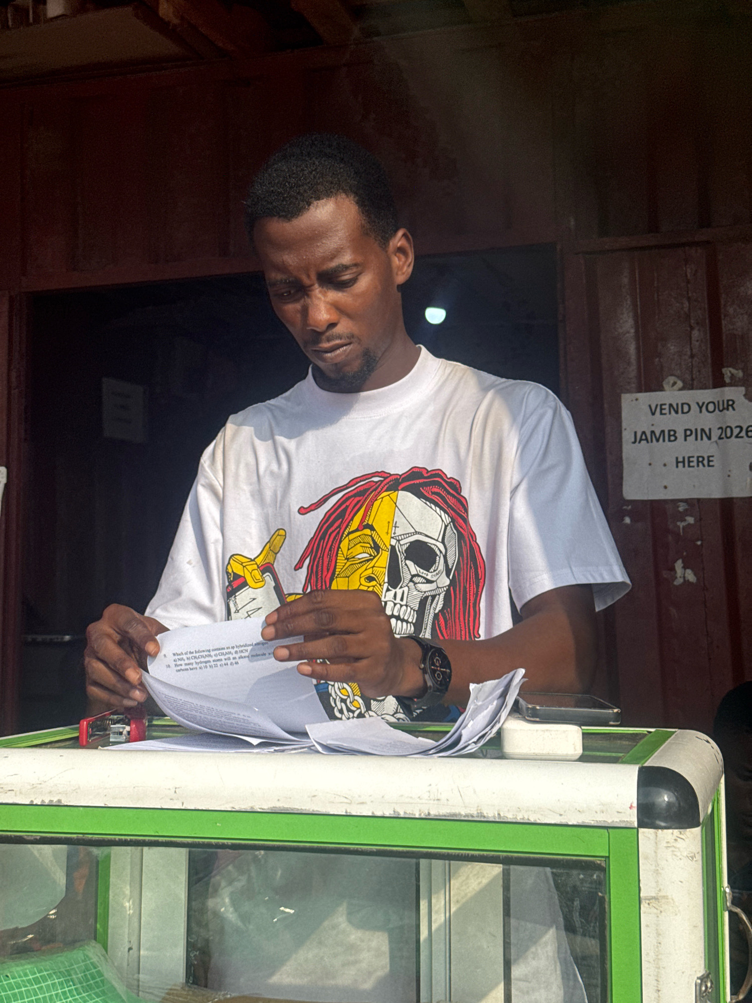 In Ihiagwa, Nigeria, a young man stands behind a white and green counter, intently reviewing documents. He is wearing a white t-shirt with a graphic design of a skull with red and yellow dreadlocks. On the counter in front of him are several sheets of paper, a red stapler, a black smartwatch, and a mobile phone. To the right of the man, a sign is affixed to a red corrugated metal surface, reading "VEND YOUR JAMB PIN 2026 HERE." The environment appears to be an outdoor kiosk or small shop.