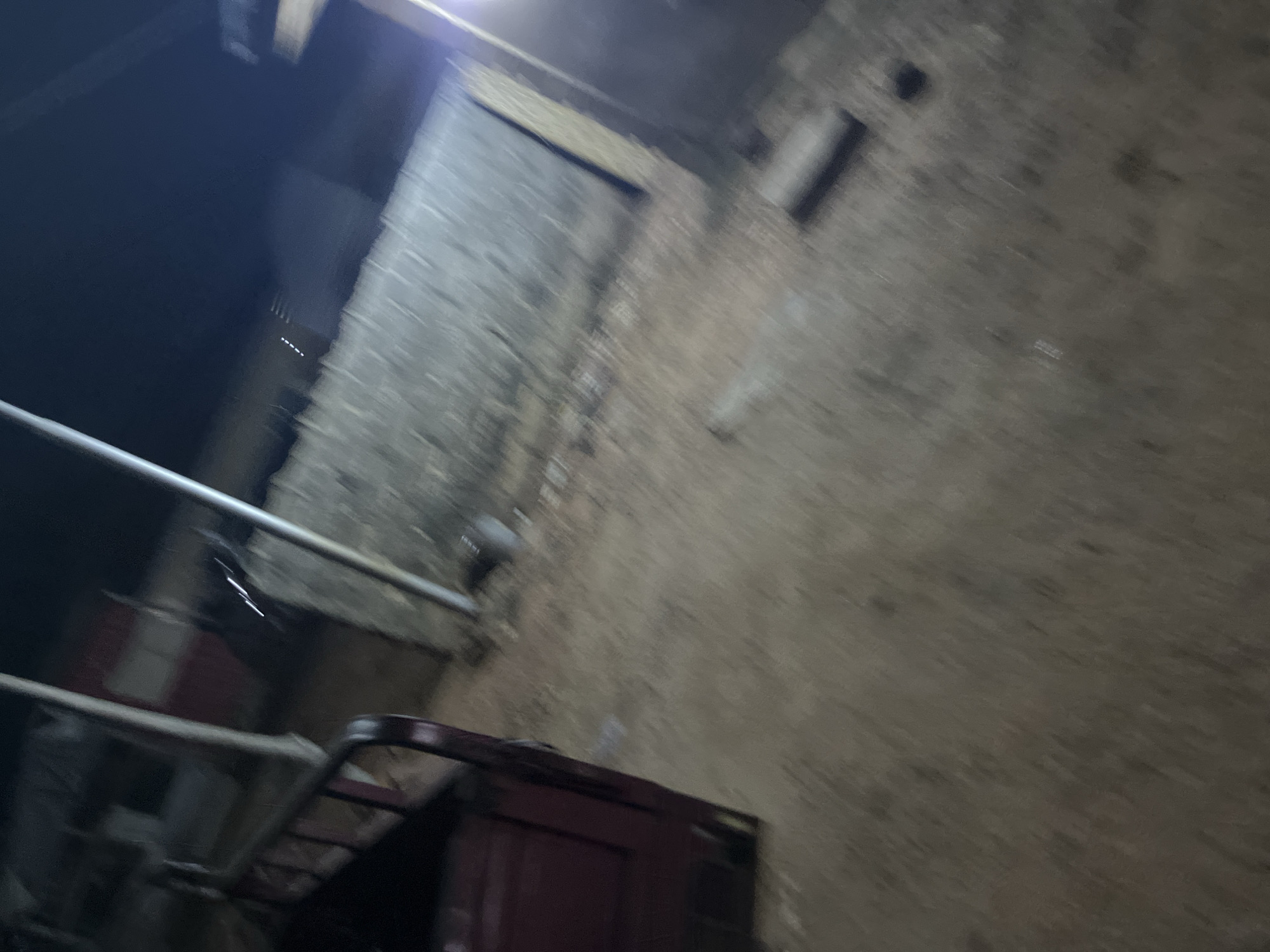 The image, taken at night in an urban environment, shows a blurry scene with a maroon tricycle on the left side. The tricycle appears to be parked on a dirt or gravel surface. In the background, there is a rough, stone or brick wall that stretches across the frame. A vertical metal pole is visible to the left of the wall, and another pole is present to the right, supporting some sort of overhead structure. The lighting is dim, with some artificial light sources casting a glow. The overall atmosphere is indistinct due to motion blur, making it difficult to discern specific details about the surrounding structures or any individuals that might be present.