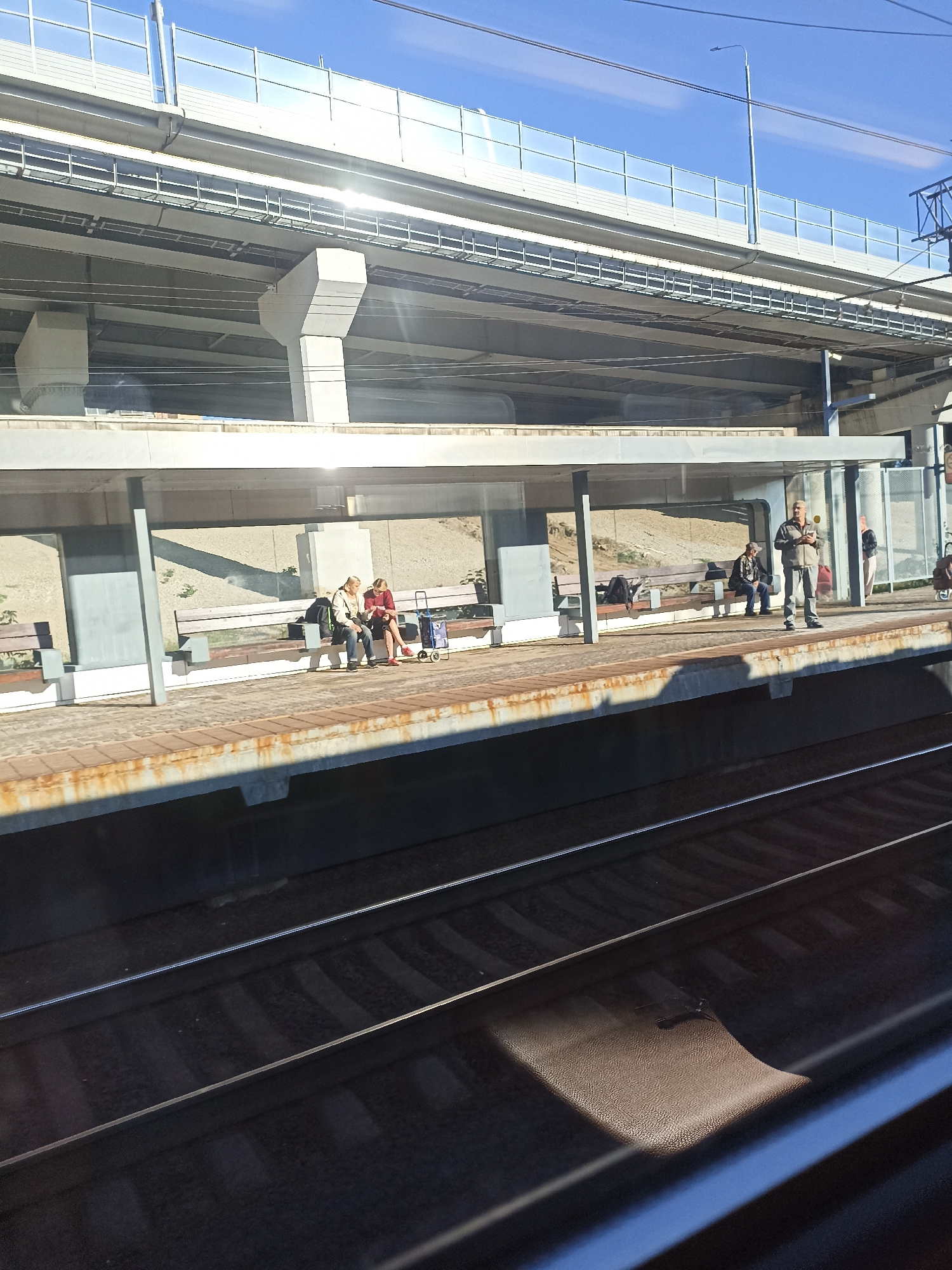 A train platform in Moscow, Russia, is depicted on a sunny day, viewed from a train window. An elevated highway structure is visible in the background, casting shadows across the platform. Several individuals are seated on benches under a covered waiting area. One person stands, holding a yellow object. The train tracks are in the foreground, with the perspective suggesting the viewer is on a moving train.