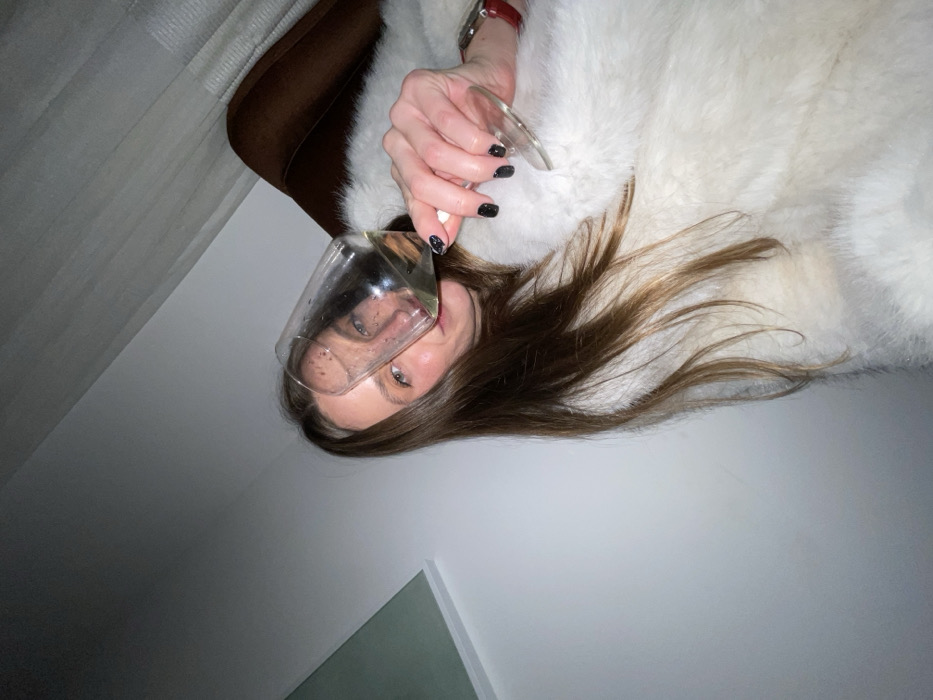 A woman lies in a white fur coat holding a glass of liquid to the camera. Part of her face is seen through the glass. She has brown hair and painted black nails. Her wrist displays a red-banded watch. A brown chair sits behind her head. The background is a light gray wall with a hint of green in the lower left corner.