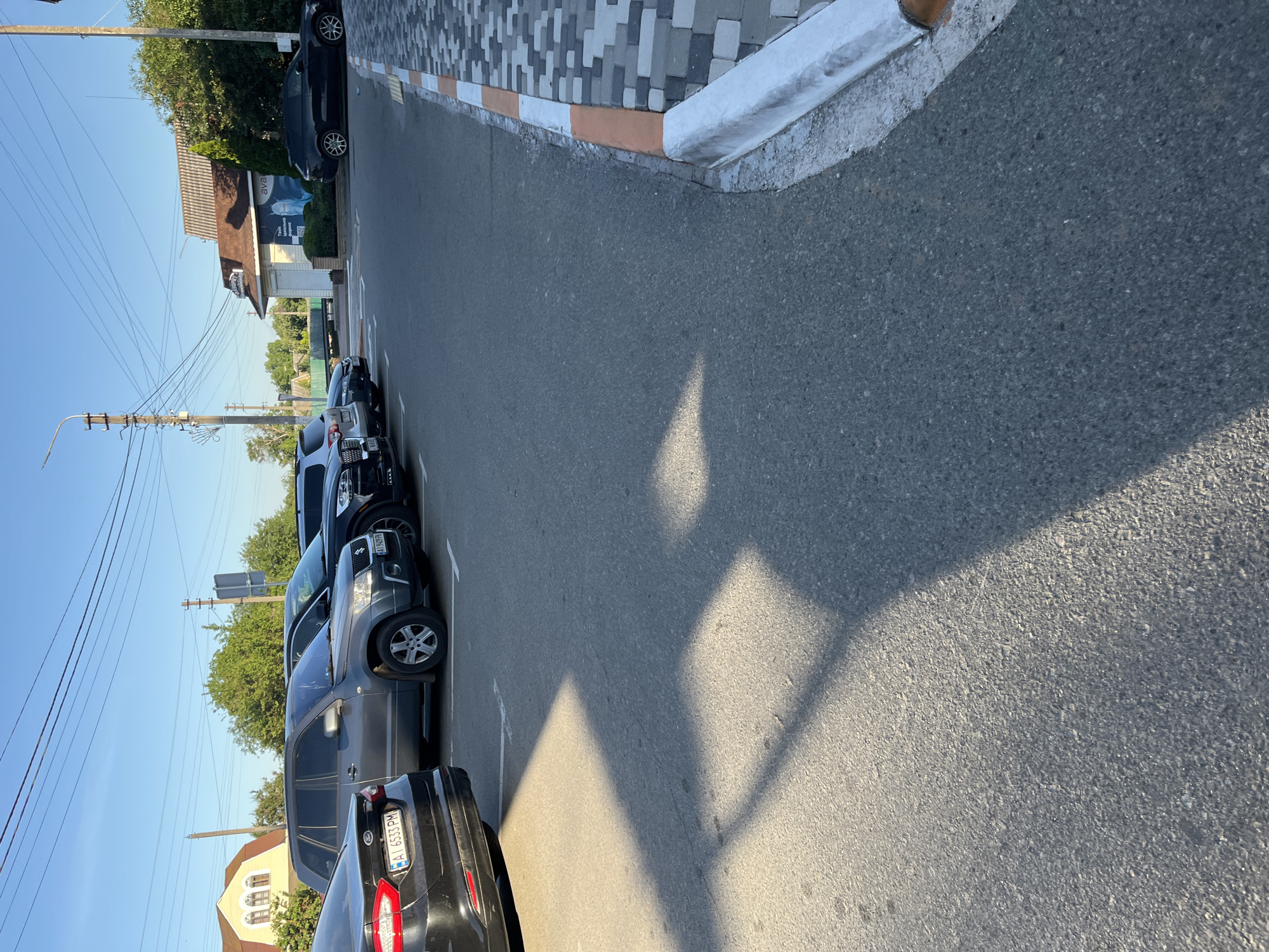 A parked car lot is shown in Kalynivka, Ukraine. The lot is paved with asphalt and bordered by a new sidewalk with orange and white trim. Several cars are parked in the lot, including a black Ford, a grey SUV, and a black SUV. The cars are parked in marked spaces. In the background, a yellow house with arched windows and a building with a sign are visible. Trees and power lines are also present in the scene. The sky is clear and blue. The lighting suggests it is late afternoon, casting long shadows across the parking lot.