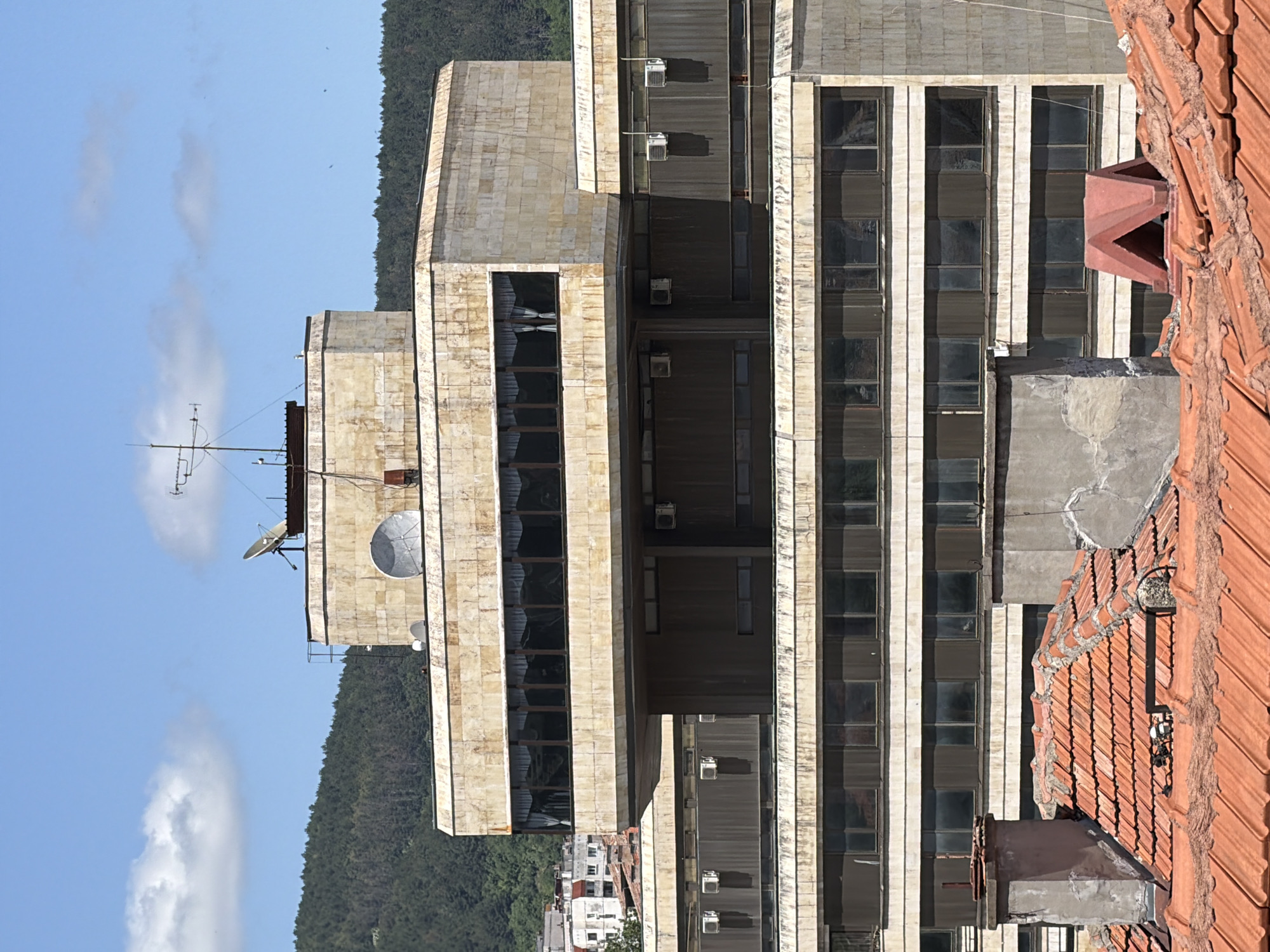 The image shows a detailed view of the Shumen Hotel in Shumen, Bulgaria, on a sunny day. The hotel is a large, multi-story building with a Brutalist architectural style, characterized by its imposing concrete structure and repetitive window patterns. The exterior walls appear to be clad in light-colored stone tiles, showing signs of age and weathering.
The topmost section of the hotel features a prominent, rectangular structure clad in similar stone tiles, with a band of dark windows running around its perimeter. On the roof of this section, a satellite dish and an antenna array are visible, suggesting communication equipment.
Below this upper section, several floors are visible, each with rows of windows. Many of these windows are dark, either due to closed blinds or the angle of the sun. Air conditioning units are mounted on some of the exterior walls. The lower foreground of the image reveals a tiled rooftop, characteristic of older buildings in the city, with chimneys and weathered concrete structures. The background shows a hillside covered in dense green forest, with a clear blue sky and scattered white clouds above. No people are visible in the image.