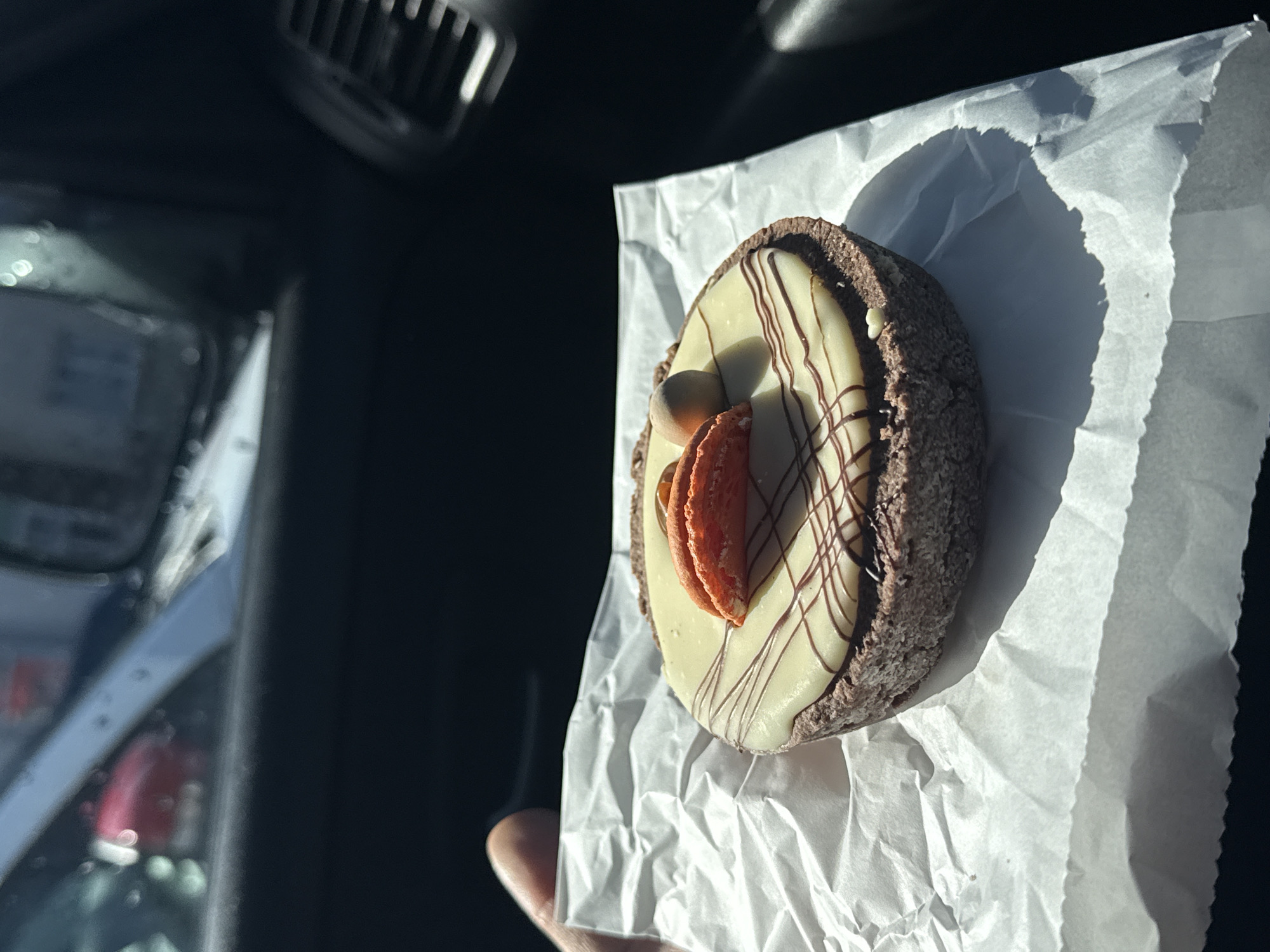 The image displays a close-up of a chocolate tart held by a human hand inside a vehicle, location Unknown, Unknown. The tart features a dark brown, textured crust and is topped with a smooth, light cream-colored icing. A pattern of thin, dark brown chocolate drizzle crosses the icing. Centered on the tart are two elongated, orange-colored confectionery pieces, possibly halves of a macaron or candied fruit, accompanied by a single, spherical, light brown nut. The tart rests on a piece of crumpled white parchment paper, supported from underneath by a visible thumb and index finger on the left. Direct sunlight illuminates the scene, casting sharp shadows on the parchment paper. The immediate background shows the car's dark interior, including a black air vent with horizontal slats on the upper right. Through the car's windshield, the blurry exterior environment is visible, featuring a light grey car pillar on the upper left, a partially obscured red vehicle, and indistinct building shapes in the distance. Water droplets are visible on the exterior of the windshield.