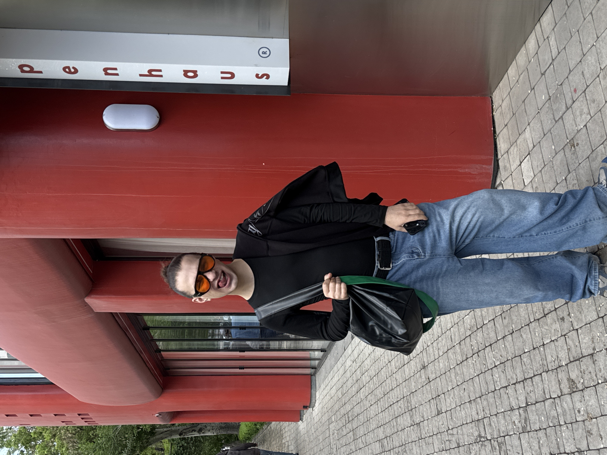 A man stands on a paved sidewalk next to a red building with the word "opus" written vertically on the side. The building appears to be of modern architectural design, with repeating red panels and visible windows. The man is wearing a black long-sleeved shirt, blue jeans, and orange-tinted sunglasses. He has a black jacket draped over his shoulder and is holding a black bag with a green strap. He is looking at the camera and smiling. The image is taken outdoors during daylight hours. This is the Cafesjian Center for the Arts in Yerevan, Armenia.