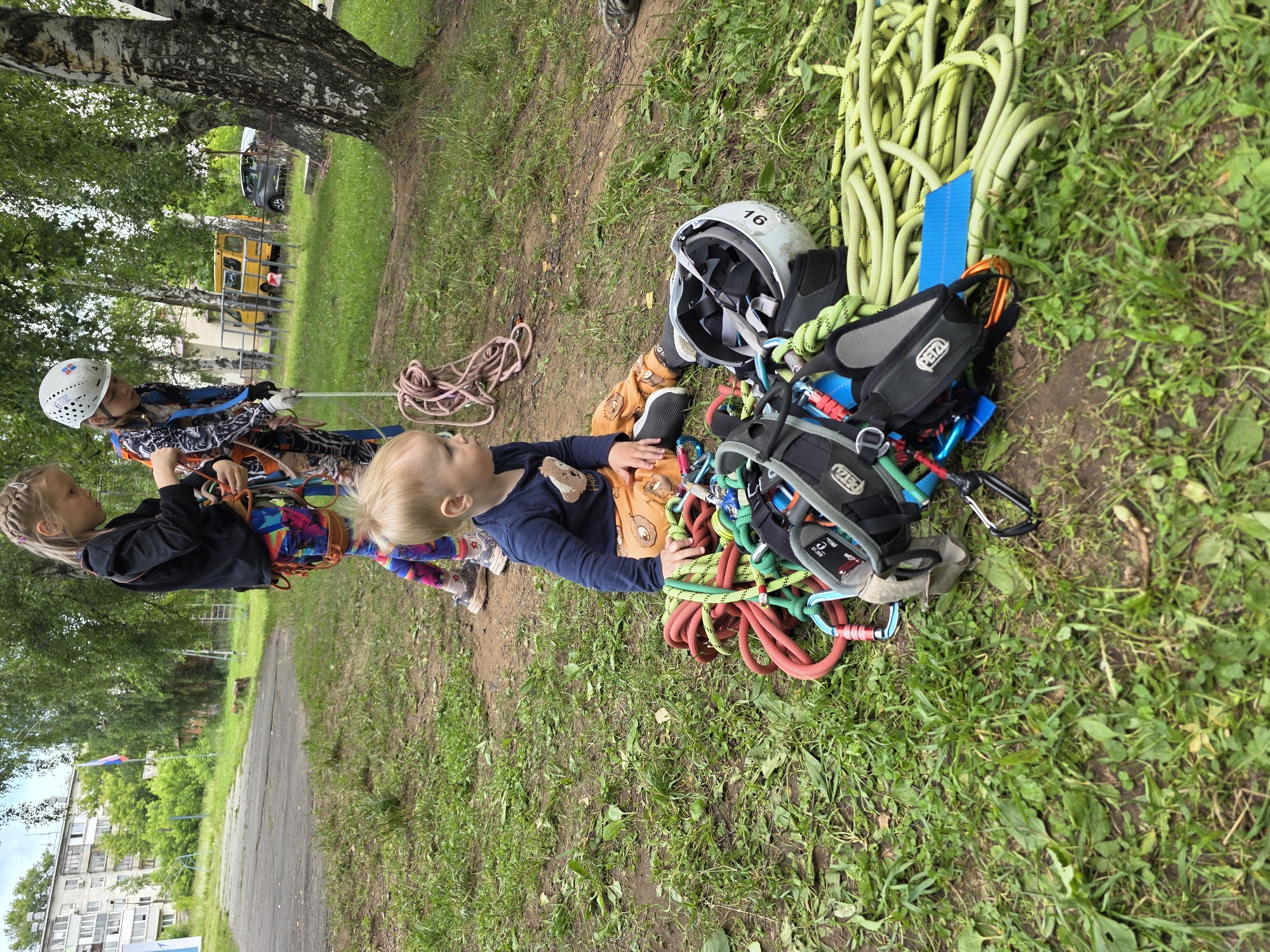 An outdoor scene shows two young girls and a toddler on a grassy area with climbing equipment, possibly in Chaikovsky, Russia. The girl on the left, with braided blonde hair, wears a black jacket and colorful leggings, and appears to be adjusting climbing gear. Beside her, a girl wearing a white helmet and a patterned suit is holding a rope. The toddler, sitting on the ground, wears a dark blue shirt with a bear design and orange pants with animal patterns. In front of the toddler is a pile of climbing equipment, including ropes of various colors, harnesses, carabiners, and a white helmet. In the background, a tall birch tree stands to the right, with a residential building and a yellow van visible in the distance. The overall setting suggests an outdoor recreational activity.