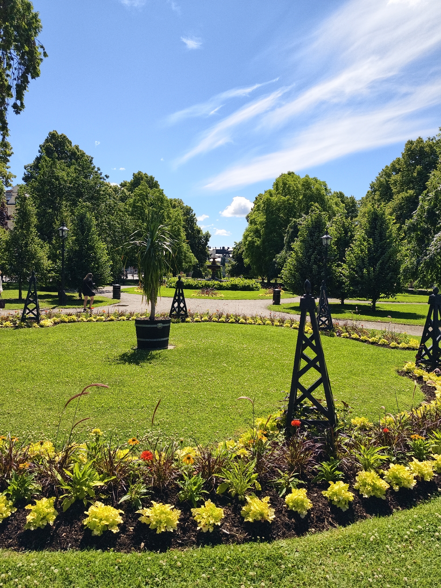 The image depicts a sunny day in a park, likely in Norrköping, Sweden, as indicated by contextual information. In the foreground, a vibrant flower bed with yellow and orange flowers lines a manicured green lawn. Several decorative black metal structures, resembling obelisks with geometric patterns, are strategically placed around the lawn and flower bed. A large potted palm-like plant stands in the center of the lawn, contained within a dark wooden barrel.
In the midground, a paved pathway winds through the park, bordered by more greenery and trees. A lamppost stands beside the path. A person wearing dark clothing is walking along the pathway, casting a shadow on the ground. A dark trash receptacle is also visible near the pathway.
The background is dominated by a dense line of mature, leafy green trees, suggesting a well-established park. Beyond the trees, parts of buildings are visible, with architectural styles that are common in European cities. The sky above is a clear blue with scattered, wispy white clouds, indicating fair weather. The overall scene conveys a sense of tranquility and natural beauty within an urban setting.