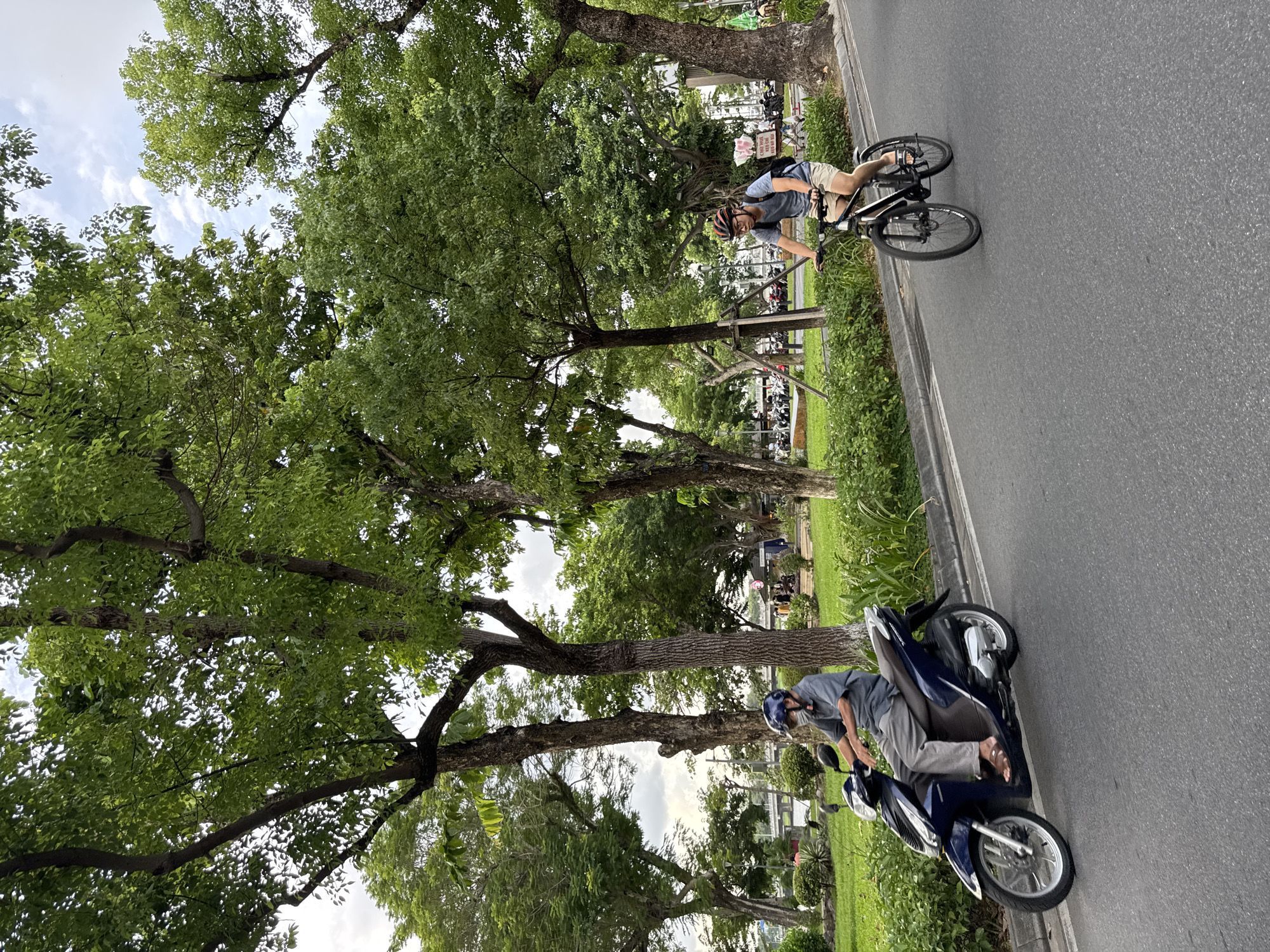 In Thuận Hóa Ward, Vietnam, a man wearing a gray shirt, gray pants, sandals, and a blue helmet rides a dark blue scooter along an asphalt road. The scooter has a silver front wheel and a silver engine casing. Beside him, a younger man with a blue t-shirt, khaki shorts, and a red helmet cycles a black bicycle with black wheels. He carries a black backpack. Both men are on a road bordered by a green lawn and lush trees with dense green foliage. The sky above is partly cloudy with white clouds against a pale blue background. In the background, more trees and a building are visible.