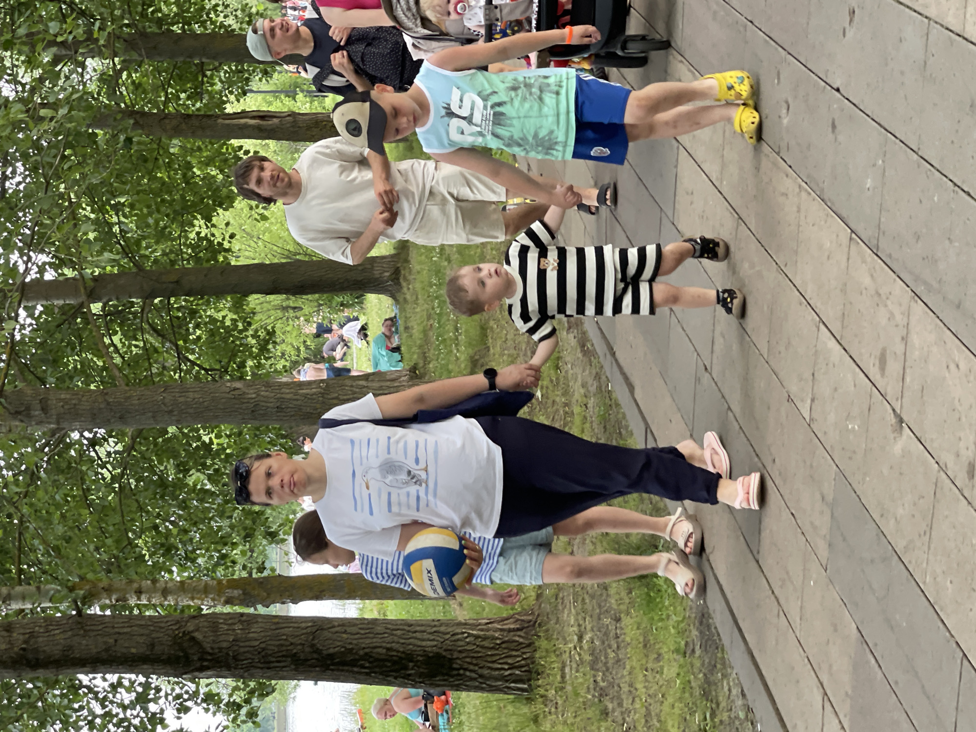 The image depicts a sunny day in a park setting, likely in Moscow, Russia, judging by the clothing styles and general atmosphere of a public gathering. A woman in the foreground, wearing dark pants and a white t-shirt with a seagull print, walks while holding a blue and yellow volleyball. To her left, a young girl in a striped shirt and shorts walks alongside her, also with a volleyball in hand. Ahead of them, two young boys are walking. The boy on the left is wearing a black and white striped outfit and is holding hands with the boy on the right, who is dressed in a light blue tank top and blue shorts, and wearing a baseball cap. A stroller is visible to the right of the boys, suggesting other individuals are present. In the background, more people are scattered throughout the park, with trees providing shade and a body of water visible in the distance. The overall scene is one of leisure and outdoor activity.