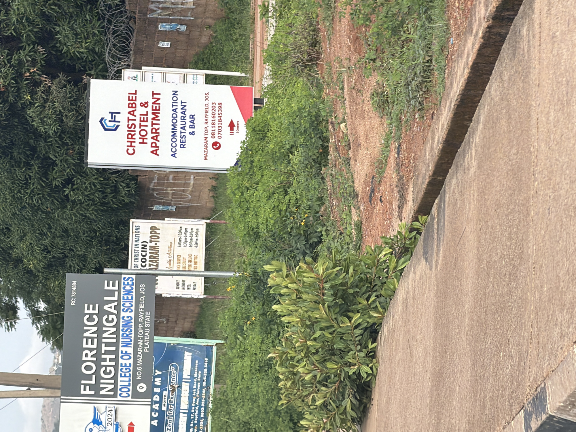 This media file captures a street view in Gura Top, Nigeria, featuring several signs advertising local establishments. The scene is dominated by lush green foliage and a concrete walkway.
The most prominent sign is for **CHRISTABEL HOTEL & APARTMENT**, which also offers **ACCOMMODATION, RESTAURANT & BAR**. This sign, displayed vertically, provides a phone number and directions. Next to it, a smaller sign indicates "10 meters".
Another significant sign is for **FLORENCE NIGHTINGALE COLLEGE OF NURSING SCIENCES**. This sign, also vertical, details the college's address on MAZARAM-TOPP, RAYFIELD, JOS, PLATEAU STATE, along with an RC number and a motto: "Zeal for Excellence". Below this, another sign appears to be for an **ACADEMY**.
A fourth sign, slightly further in the background, appears to be for **OF CHRIST IN NATIONS (COCIN)** and lists **AZARAM-TOPP** with "Weekly Activities" such as church service, Bible study, and prayer meetings, along with their timings.
The overall setting suggests an outdoor environment, possibly during daylight hours, given the clarity of the signs and the shadows. The presence of multiple signs indicates a commercial or educational area within the community. The focus is on the information presented on these signs, advertising services and institutions in the specified Nigerian location.
