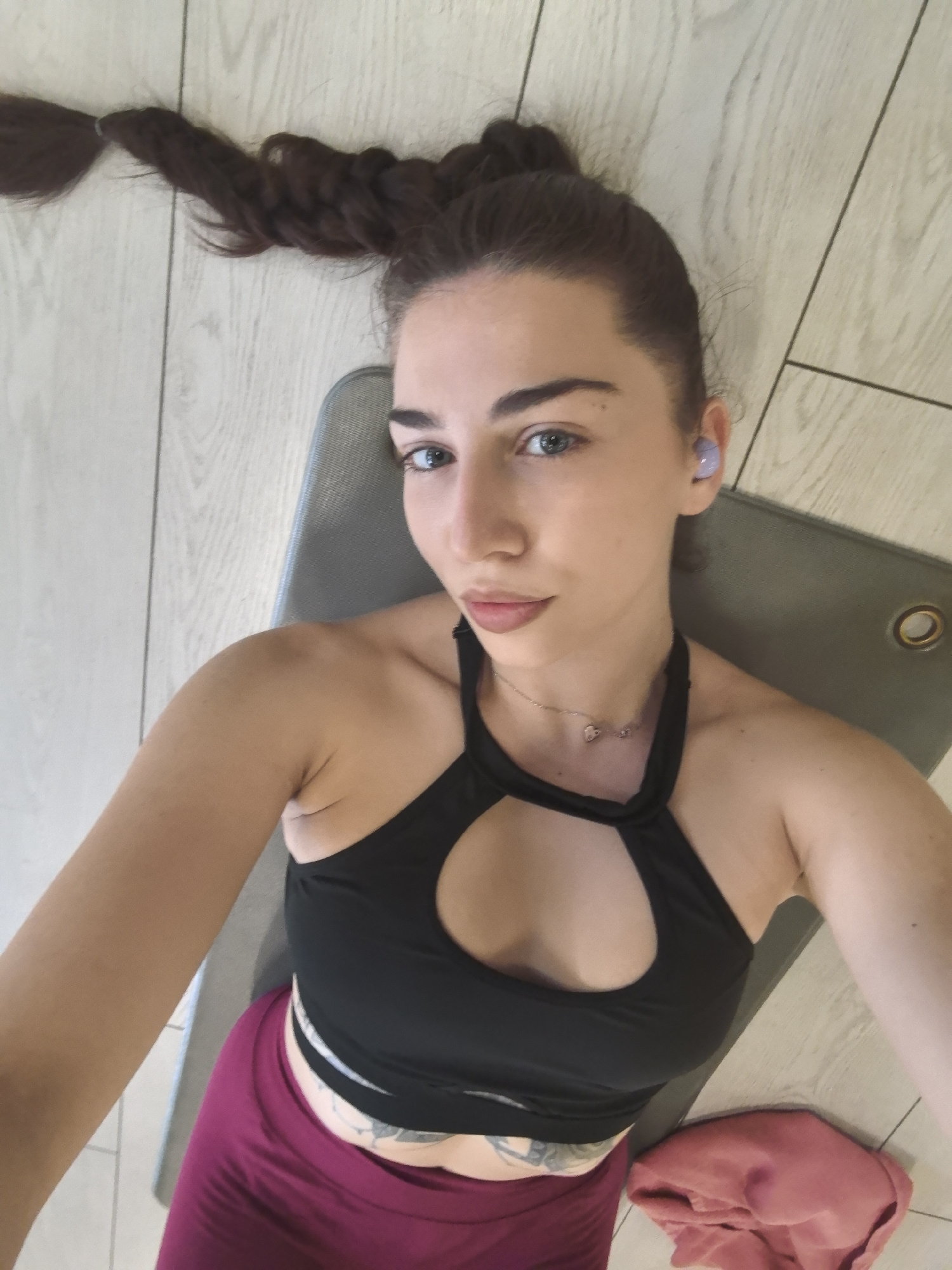 A woman is depicted lying on her back on a grey yoga mat, positioned on a light-colored wooden floor. She is looking directly at the camera, capturing a selfie from above. Her dark brown hair is styled in a long, thick braid that extends upwards and to the left from her head. She has light-colored eyes, possibly blue or green, and a calm expression.
She is dressed in athletic wear, consisting of a black sports bra with a keyhole cutout design on the chest and dark pink or magenta-colored leggings. A delicate silver necklace with small charms adorns her neck. A light purple wireless earbud is visible in her right ear. A tattoo can be seen on her lower abdomen or hip area, just below the edge of her sports bra.
Beside the yoga mat, on the wooden floor to her right, lies a folded pink or reddish towel or piece of clothing. The setting appears to be indoors, likely during the daytime, with bright and even lighting. The scene suggests a moment of rest or a break from a workout. This image is set in Stara Zagora, Bulgaria. No text is visible in the frame.