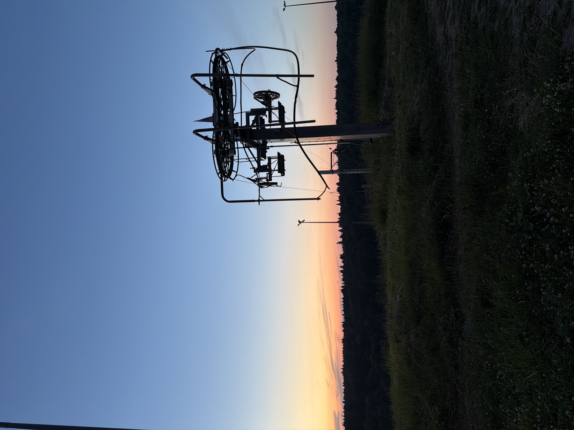 The image depicts a ski lift station silhouetted against a gradient sky during twilight. The sky transitions from a deep blue at the top to hues of orange and yellow near the horizon. A dense forest forms a dark silhouette below the horizon. In the foreground, a grassy hill is visible, with a ski lift tower prominently featured on the right side of the frame. The ski lift tower is intricate, with visible mechanical components and suspended chairs. Several poles, possibly lighting or part of the ski lift infrastructure, are scattered throughout the scene. The overall atmosphere is serene and slightly melancholic, characteristic of dusk. The location is identified as Krasnogorsk, Russia. There are no people present in the image.
