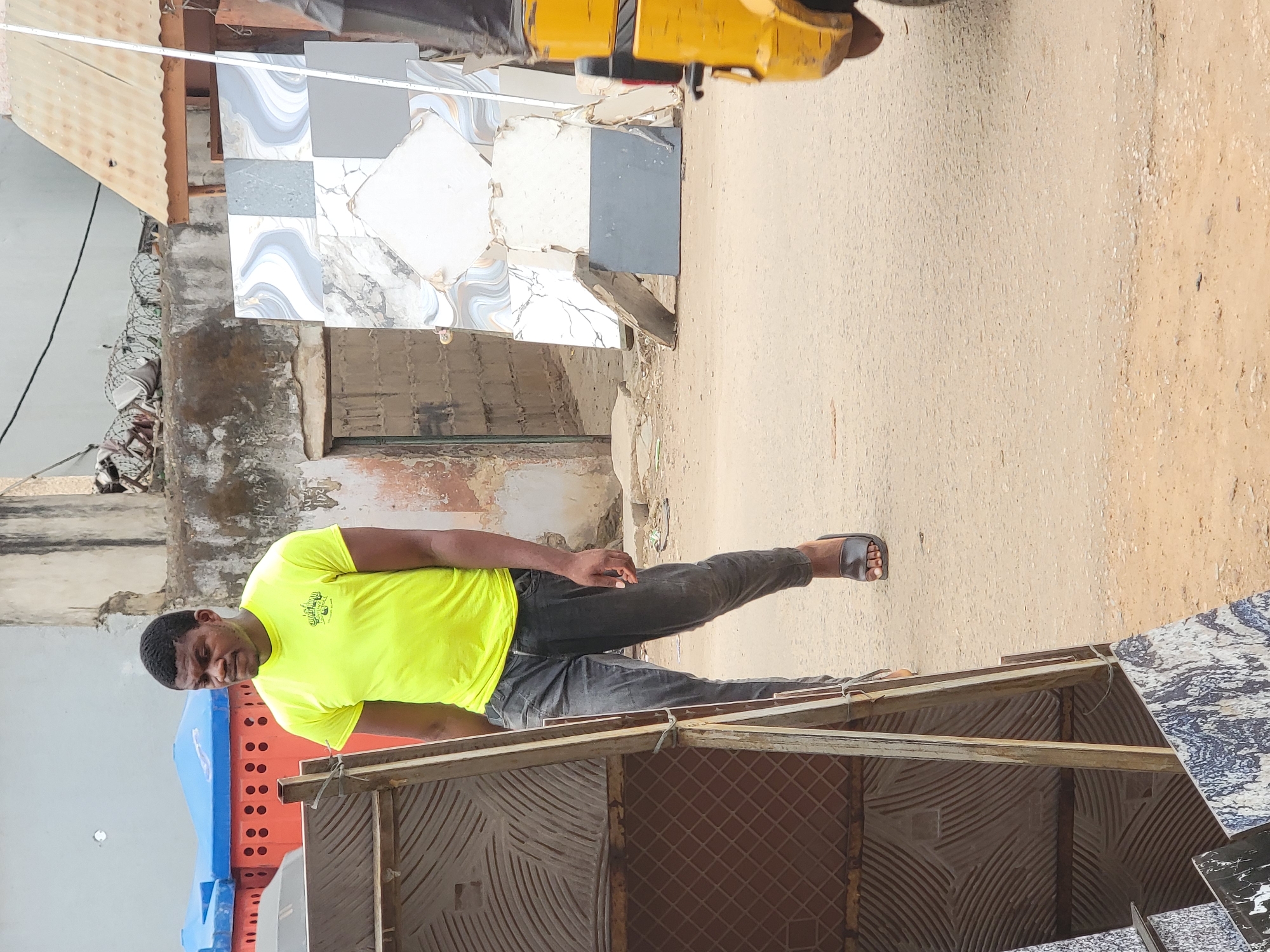 A young man stands beside a display of ceramic tiles in an outdoor setting in Maiburiji, Nigeria. The man, with dark skin, wears a bright neon yellow t-shirt, dark grey pants, and black slip-on sandals. His t-shirt features a dark graphic with the legible text "Living the DREAM". He stands with a calm, slightly smiling expression, looking towards the viewer.
In the foreground and extending into the midground, a rustic wooden structure serves as a display stand for an assortment of ceramic tiles. These tiles showcase various patterns, including marbled designs in grey and white, wavy blue and white patterns, and plain dark grey squares. More tiles are stacked or leaning in the background.
The ground is unpaved, a light brown dirt or sandy surface, consistent with a street or market area. Behind the tile display and the man, a weathered wall suggests an older building. Further in the background, a light brown corrugated metal roof is visible, along with stacks of blue and orange plastic crates. A yellow vehicle, possibly a tricycle, is partially visible in the upper background. The scene is brightly lit, indicating daytime, with soft lighting suggesting an overcast sky or indirect sunlight.