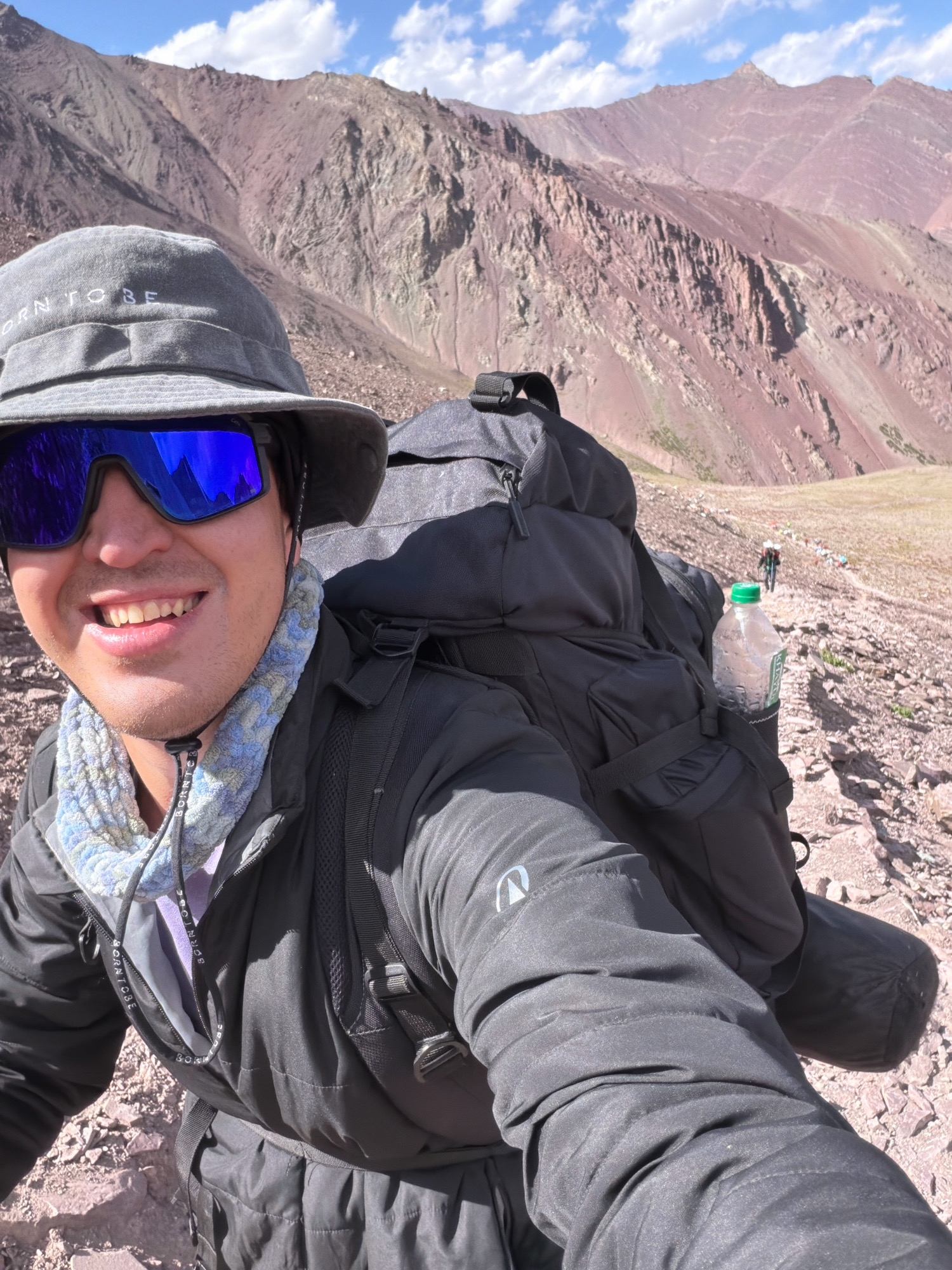 A man wearing a grey bucket hat, blue mirrored sunglasses, and a black jacket smiles for a selfie while hiking in a mountainous, rocky terrain under a partly cloudy sky. He has a large black backpack on, with a water bottle in a side pocket. A group of hikers can be seen in the distance on a winding trail. The mountains in the background are steep and a reddish-brown color. The text "BORN TO BE" is visible on his hat and on the drawstrings of his jacket.