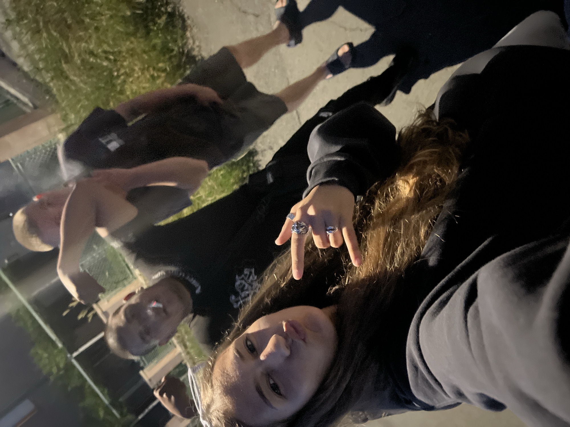 This image, captured at night or late dusk in Debelt, Bulgaria, depicts three individuals, appearing to be young adults or teenagers, posing for a selfie on what appears to be a paved surface next to an overgrown grassy area.
In the foreground, a female subject with long, wavy brown hair, wearing a dark long-sleeved top or hoodie, holds the camera for the photograph. Her lips are pursed in a pout, and white-framed sunglasses rest on top of her head. Her left hand is raised, forming a 'corna' or 'rock on' gesture, with multiple rings visible on her fingers: a large ornate silver-colored ring on her index finger and two smaller silver-colored rings on her middle finger.
Directly behind her and slightly to the left, a male subject with short, dark hair wears a dark t-shirt with a white graphic or text on the chest, and a silver-colored chain around his neck. He holds a lit cigarette between his lips, and his right arm is bent with the bicep visibly tensed. His left hand is seen grasping the bicep of the third male subject.
The third male subject is positioned furthest back and to the right, mostly visible from the waist up. He has short, light-colored hair and wears a dark t-shirt and dark shorts. His left bicep is being held by the second male subject. He wears dark slides or sandals on his feet.
The environment is dimly lit by an unseen artificial light source, casting shadows. The ground consists of a paved surface adjacent to a strip of unkempt grass and weeds. In the background, vague outlines of structures, possibly buildings or fences (a chain-link fence is discernible behind the smoking male), and dark foliage are visible.