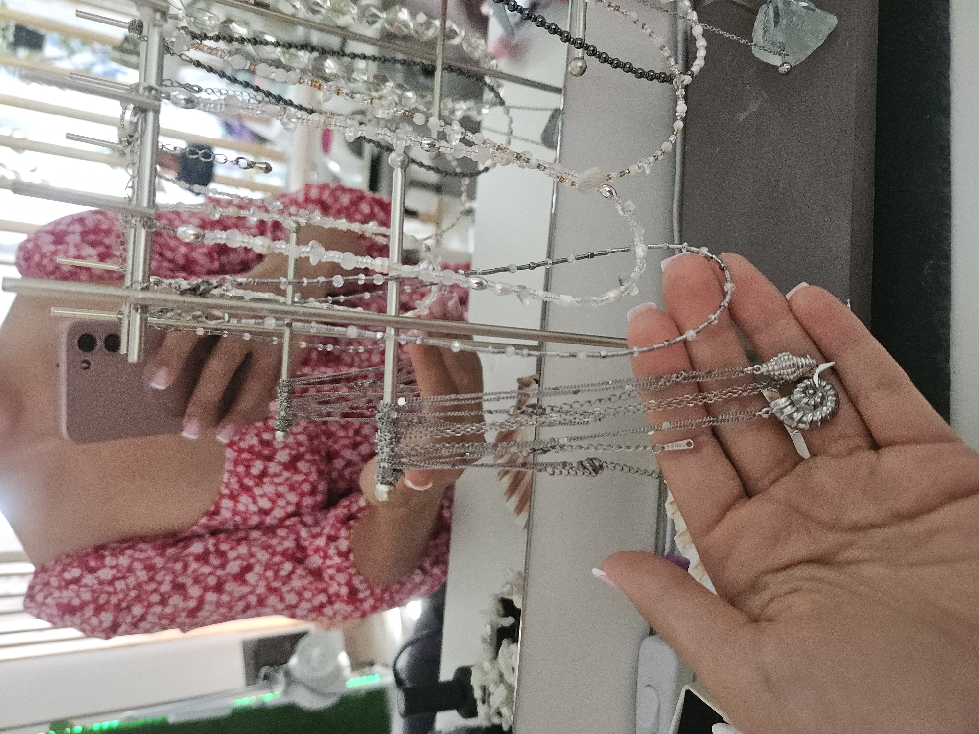 A person is holding a collection of necklaces in their hand. The necklaces are displayed on a tiered rack, with various beads and chains visible. The person is wearing a red and white floral patterned dress. The reflection in a mirror shows the person holding a pink smartphone. The scene appears to be indoors, with natural light coming from a window with blinds. Some greenery and furniture are visible in the background reflection. The focus is on the hand and the necklaces. A small tag on one of the necklaces appears to have the text "sensi" or a similar word.