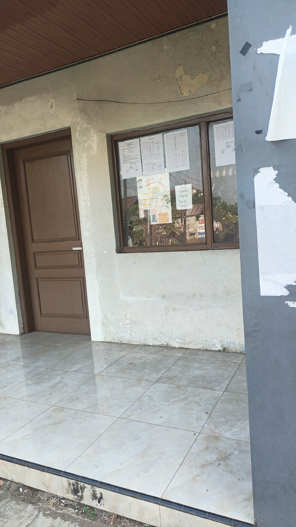 The image shows the exterior of a building. A brown wooden door is on the left side of the image. To the right of the door is a window with papers posted inside. The wall is a light color and appears to be somewhat weathered. The ground in front of the building is tiled with light-colored square tiles. In the background, through the window, reflections of trees and buildings can be seen. On the right side of the image, a dark grey vertical surface is partially visible. The scene is likely located in Margorejo, Indonesia, given the context.