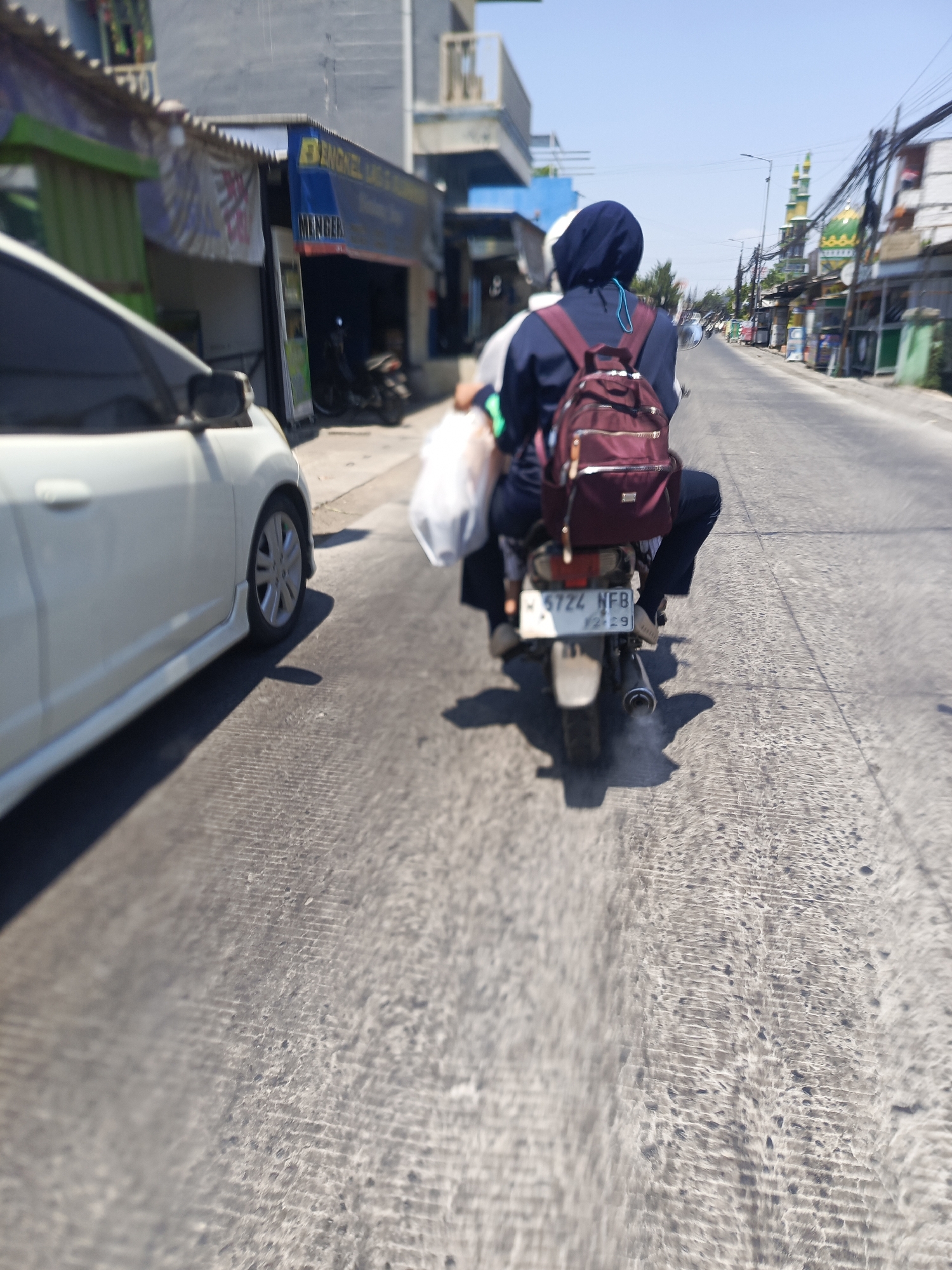 In Kwangsan, Indonesia, on a sunny day, a white car is parked on the side of a paved road. A person wearing a dark blue hijab and a maroon backpack is riding a motorcycle, with another person in front of them. The motorcycle has a license plate that reads "6724 NFB 12-29". Buildings line the road, and power lines stretch overhead. The road surface is textured with parallel lines and scattered dark spots, indicating wear and tear. A building with a blue awning and the text "ENGKEL JAG..." is visible on the left side of the image. In the distance, a mosque with green domes and minarets stands against the clear blue sky. The scene suggests a typical day in an Indonesian town, with traffic, businesses, and religious buildings coexisting along the street.