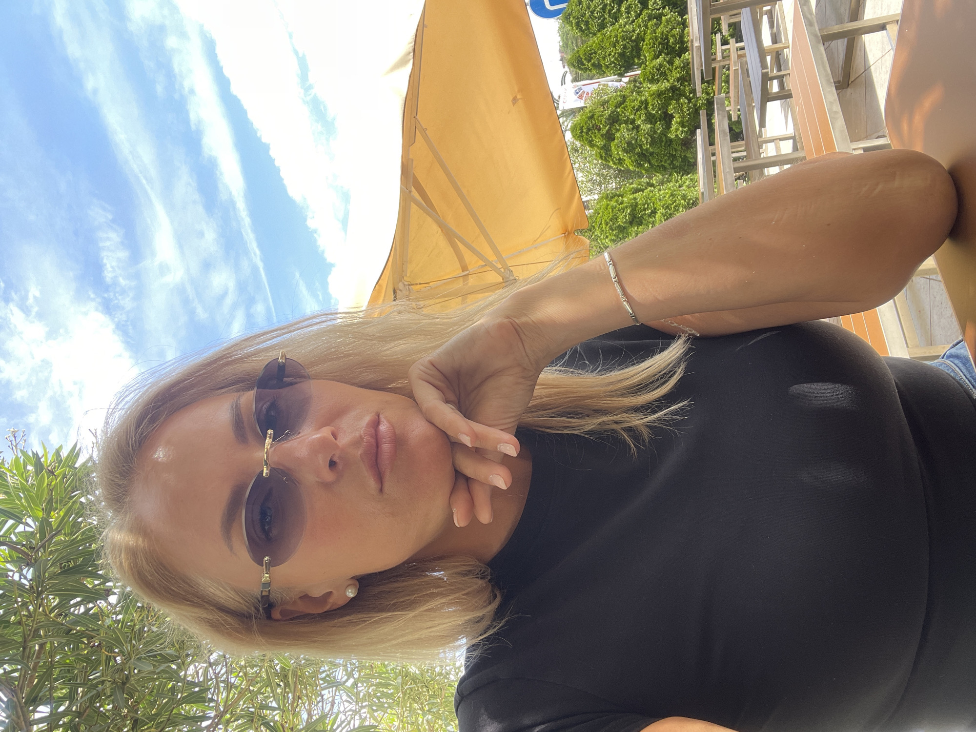 A woman with blonde hair and sunglasses is posing for a selfie outdoors under a yellow umbrella on a sunny day. She is wearing a black top and has a silver bracelet on her left wrist. Her right hand is resting on her chin. The background includes green trees, a blue sky with clouds, and outdoor furniture. This scene appears to be taken in a public area, possibly a cafe or park in Sochi, Russia.