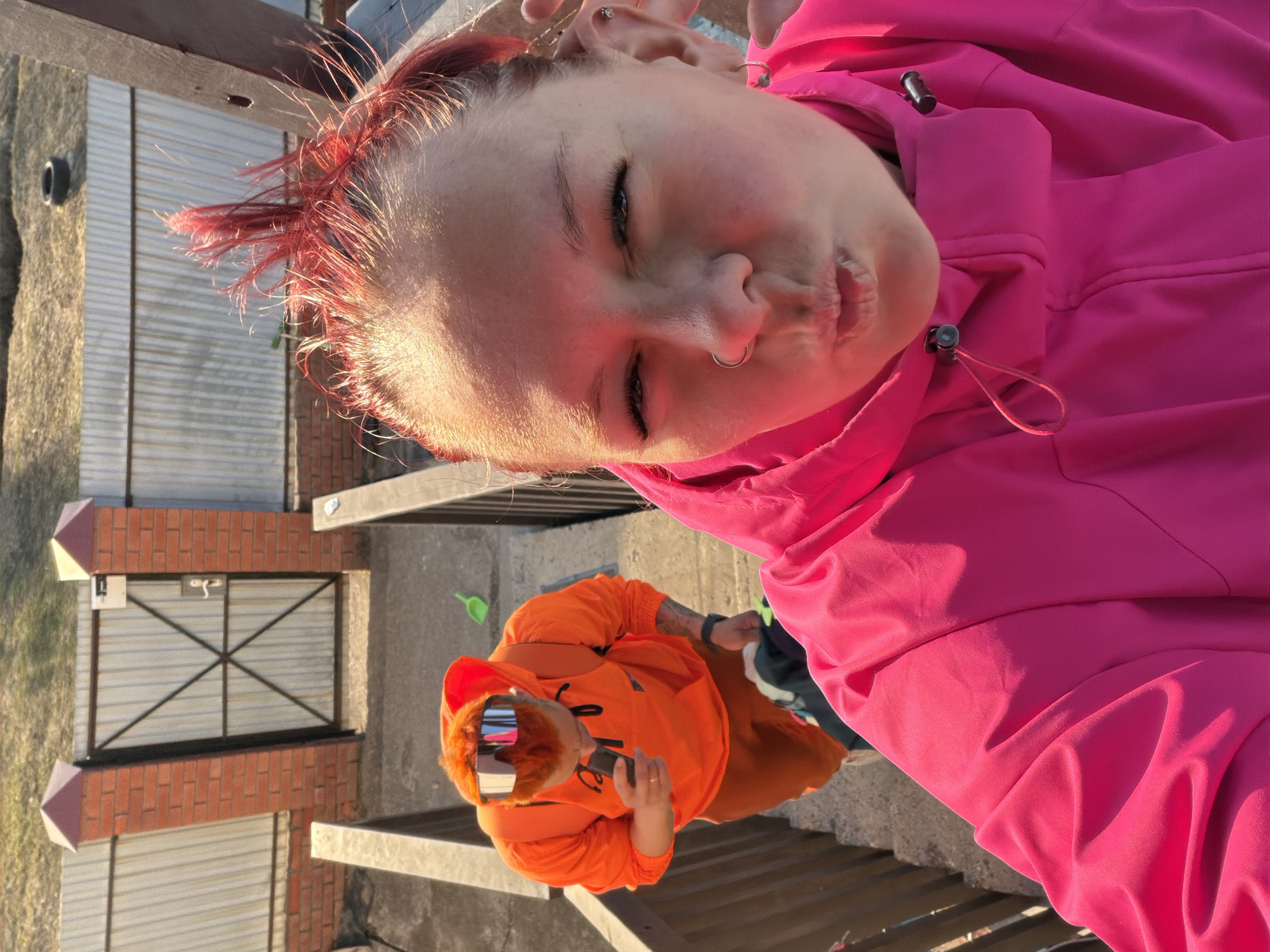 A photograph shows two people. One person, wearing a bright pink hooded jacket, is oriented at the top of the frame. The person has fair skin, short red hair, and pierced nose and ears. The second person is wearing a bright orange hooded jacket and mirrored sunglasses. They are holding a cell phone in their right hand. The setting appears to be outdoors, next to a brick structure, which is likely a fence or a small building. The photograph was likely taken near Chaikovsky, Russia. The specific location within the city cannot be determined from the image.
