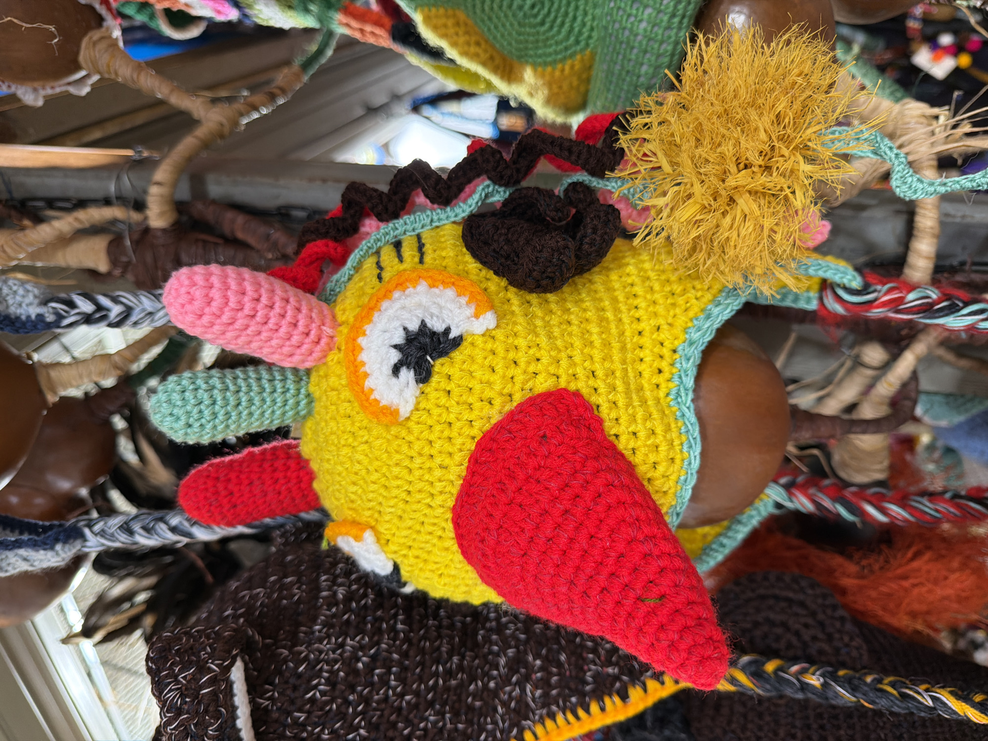 This is a close-up, eye-level shot of a brightly colored, crocheted hat shaped like a bird's head, likely displayed for sale in an outdoor market in Nha Trang Ward, Vietnam. The hat is predominantly yellow, with a large red beak, and white and orange eyes outlined in black. Green and pink crocheted elements protrude from the top of the head, resembling ears or feathers. A textured brown crocheted piece and a thin, zig-zagging light blue crocheted strand are visible on the side of the head. A fluffy, yellow, fringe-like attachment is prominent on the right side. Multiple braided cords, in various color combinations of red, white, black, and yellow, hang from the hat, suggesting it is designed to be worn. The background is blurred, but appears to be a collection of similar handcrafted items, possibly other hats or decorations, made of yarn, gourd-like materials, and natural fibers, suggesting a vibrant marketplace setting. The lighting is natural and even, indicating an outdoor or well-lit indoor environment.