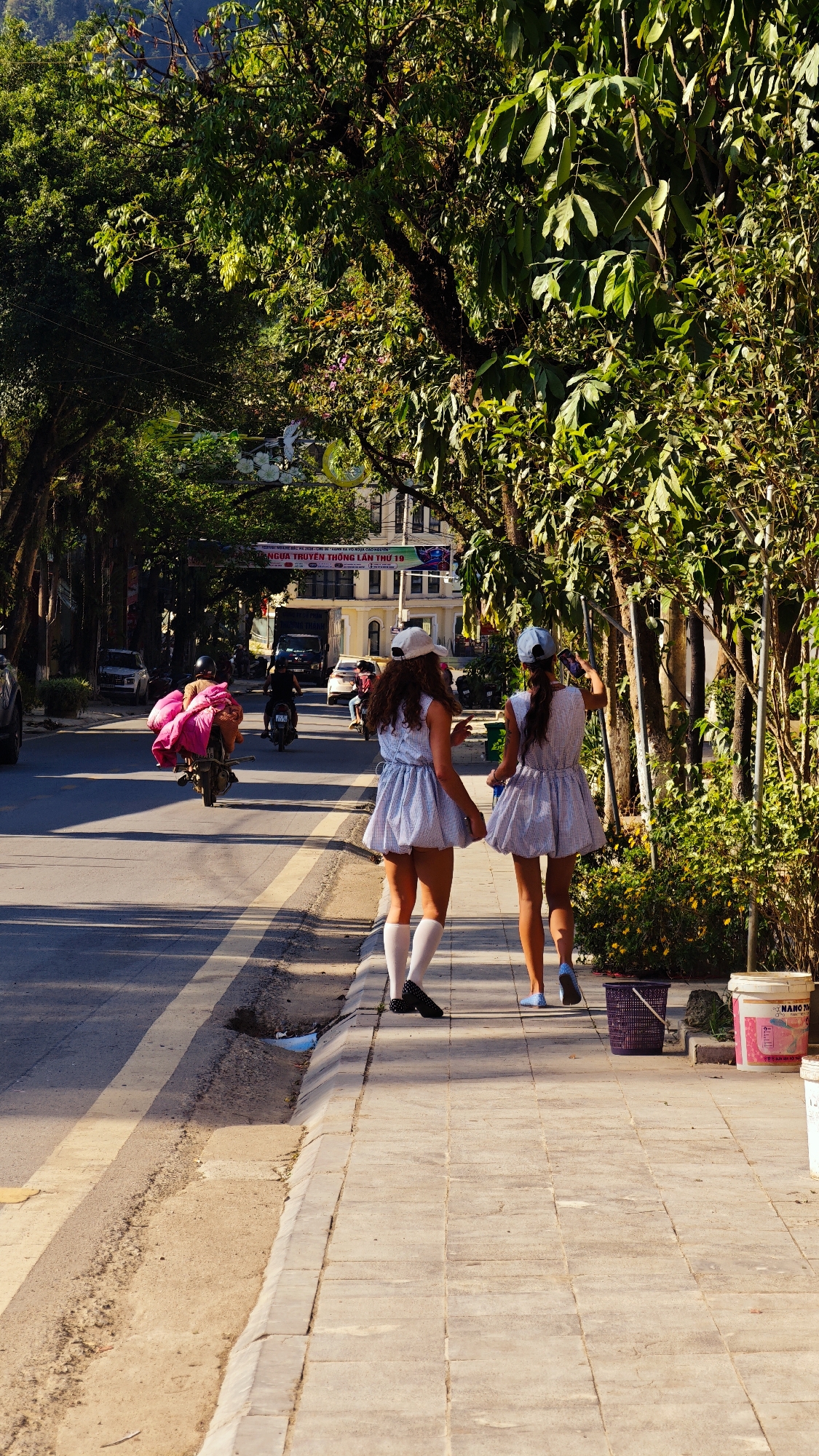 Two young women dressed in matching blue and white dresses and baseball caps walk along a paved sidewalk. One wears white knee-high socks and black shoes; the other wears blue slip-ons. They are walking away from the viewer.
The sidewalk is adjacent to a road with a yellow dividing line. Motorbikes and vehicles travel along the road. One motorbike carries a large, pink load. Overhead, trees form a canopy over the road.
In the background, a multi-story building is visible, along with a banner written in Vietnamese. Buckets and a plastic basket sit on the sidewalk near the women. The location is Bắc Hà, Vietnam.