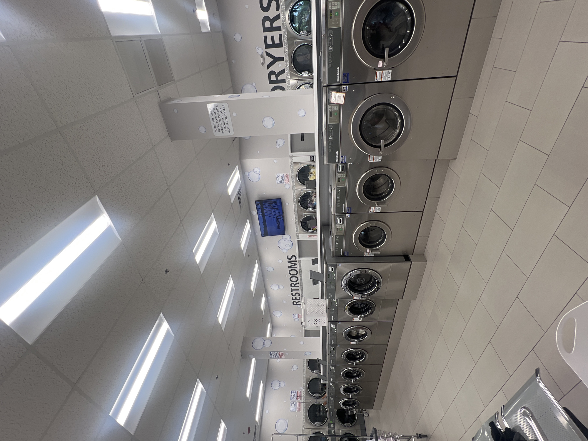 This is an indoor shot, likely of a laundromat, with rows of large, industrial-looking washing machines lined up against a tiled wall. Above the machines, "DRYERS" is written in large letters on the wall, surrounded by decorative bubbles. There's a pillar or wall feature with "RESTROOMS" written vertically, also decorated with bubbles, leading towards the back of the laundromat. A sign on the pillar indicates items not allowed in the washers or dryers. The ceilings are tiled with embedded fluorescent lights. Seating is visible on the right side of the shot. The location appears to be in an urban area, though the specific city and state are unknown.