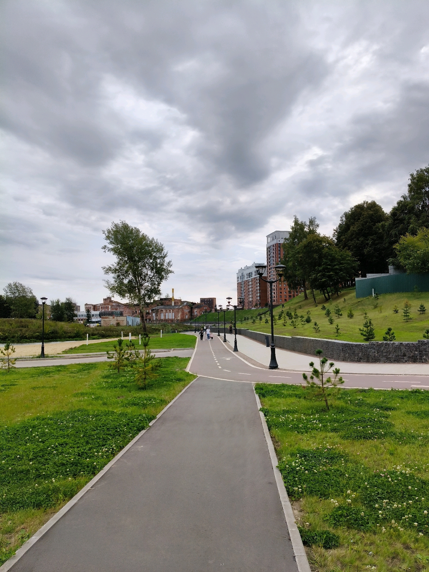 The image depicts a paved walkway and a cycling path running parallel through a green park in Naro-Fominsk, Russia. The walkway is bordered by grass and some small trees. In the background, there are residential buildings and older industrial structures. The sky is overcast with gray clouds. Several people are walking on the path.