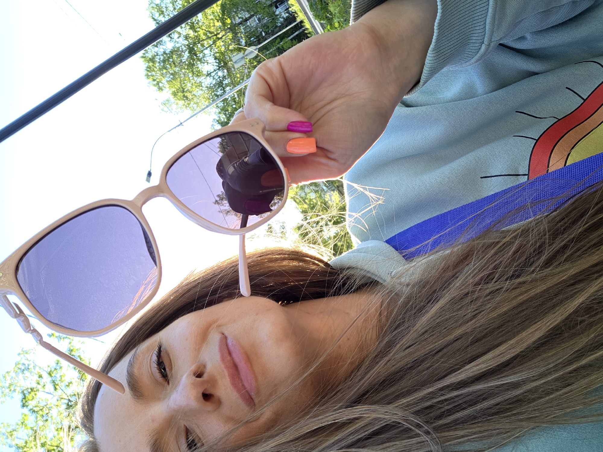 The image shows a woman in an outdoor setting, possibly in Birobidzhan, Russia, given the instructions. The woman is holding sunglasses in her right hand, with two of her fingernails painted bright pink and orange, respectively. The sunglasses have a light pink frame and purple lenses. She is wearing a light blue sweatshirt with a rainbow design on the sleeve. A blue strap is visible across her chest. Her hair is light brown with blonde highlights. Trees and a sky are visible in the background.