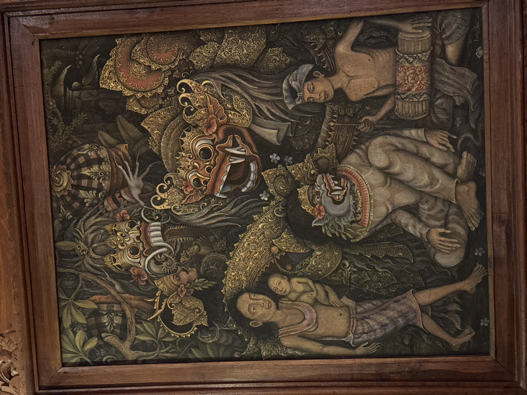This intricate, framed painting depicts a complex narrative scene, likely drawn from Balinese mythology or folklore, characteristic of traditional Balinese art.
**Subjects:** The artwork features numerous interwoven figures against a dark, densely patterned background. Prominent are several human-like figures in various states of repose or activity, some adorned with elaborate headwear or clothing. In the upper part, a figure lies nude from the waist up, holding what appears to be a drum. Nearby, another figure with long dark hair, wearing a light blue head covering, appears to be resting. Towards the bottom, a woman lies with a baby resting on her chest, both looking upwards. Interspersed among these human figures are larger, more monstrous or divine entities, characterized by elaborate, stylized masks. A large red mask with fangs and wide eyes dominates the left-center, while another formidable mask, primarily white and gold, is seen to its right. The background is filled with dense, dark foliage and intricate, swirling golden and reddish patterns that resemble decorative carvings or natural elements. The painting is enclosed within a dark wooden frame with carved details.
**Activity or Event:** The scene suggests a dramatic, perhaps mythological, event. Some figures appear to be resting or deceased, while others, particularly those associated with the masks, convey power and intensity. A figure in the upper right is playing a stringed instrument, contributing to a sense of a ritual or ceremonial atmosphere. The intertwining of figures and patterns creates a dynamic and immersive narrative.
**Notable Details:** The art style is highly ornate, utilizing rich earth tones, dark greens, and striking gold and red accents against a deep black or dark brown background. The gold details often highlight decorative elements on clothing, headdresses, and the masks, giving the impression of gilded surfaces. The masks strongly resemble Balinese Barong or Rangda figures, indicating a spiritual or mythological context. The intricate patterns throughout the background create a mystical forest or jungle setting. The overall mood is intense and captivating, blending elements of life, death, and supernatural presence.
**Visible Text:** In the upper right corner of the painting, a signature "I. Lub. Badra" is visible, likely identifying the artist.