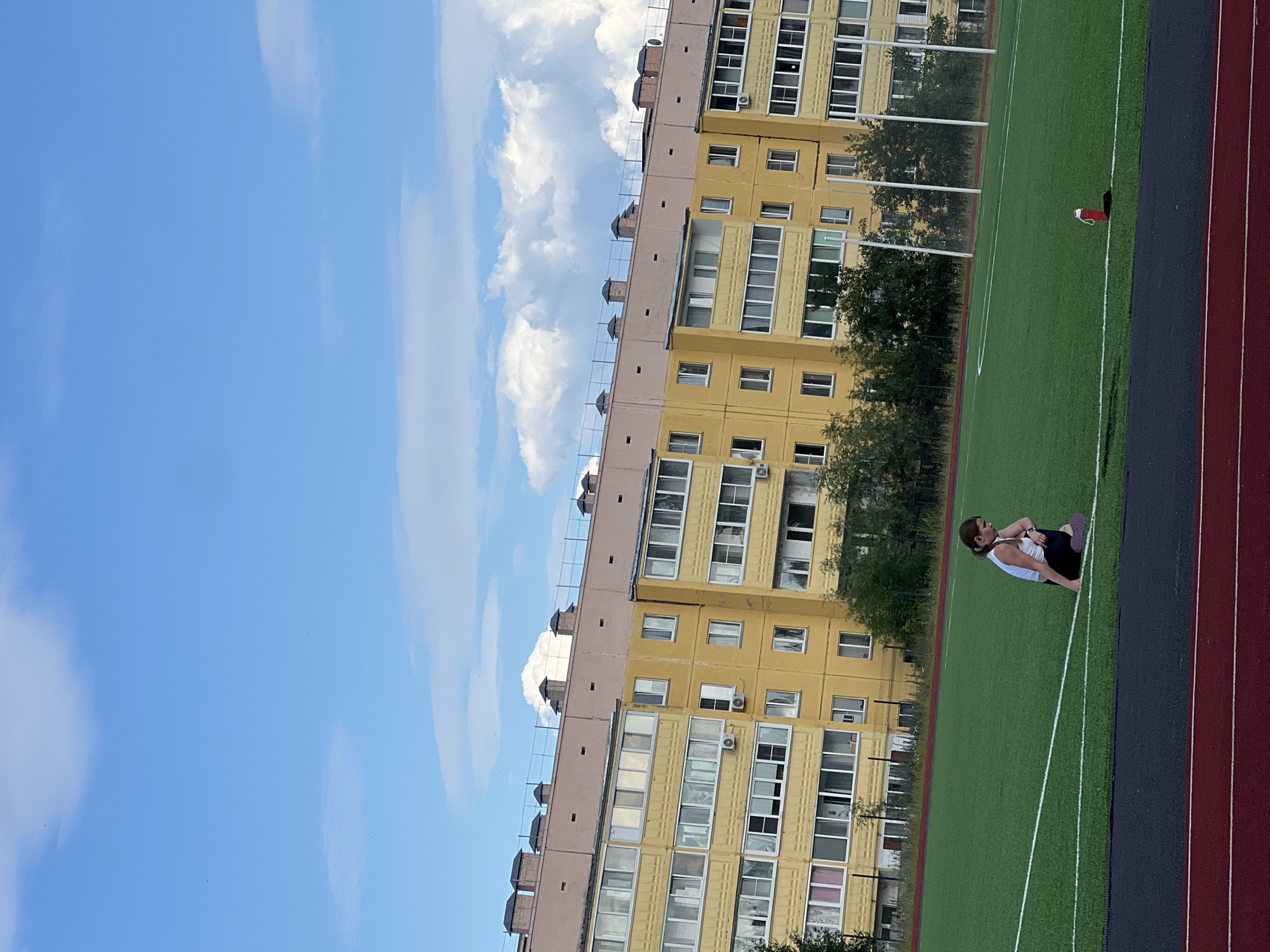 The image depicts a young woman with brown hair, wearing a white tank top and black athletic shorts, kneeling on a bright green artificial turf field. She is positioned on the left side of the frame, facing right with her head tilted down. A purple mat is visible beneath her knees. The field is marked with white lines, and a red water bottle is lying on its side near the right side of the frame. In the background, a long, yellow, multi-story apartment building stands under a blue sky with scattered white clouds. The building has numerous windows and balconies, and several trees are visible in front of it. The foreground shows a dark red running track. The scene appears to be a sports facility in Noviy, Russia.