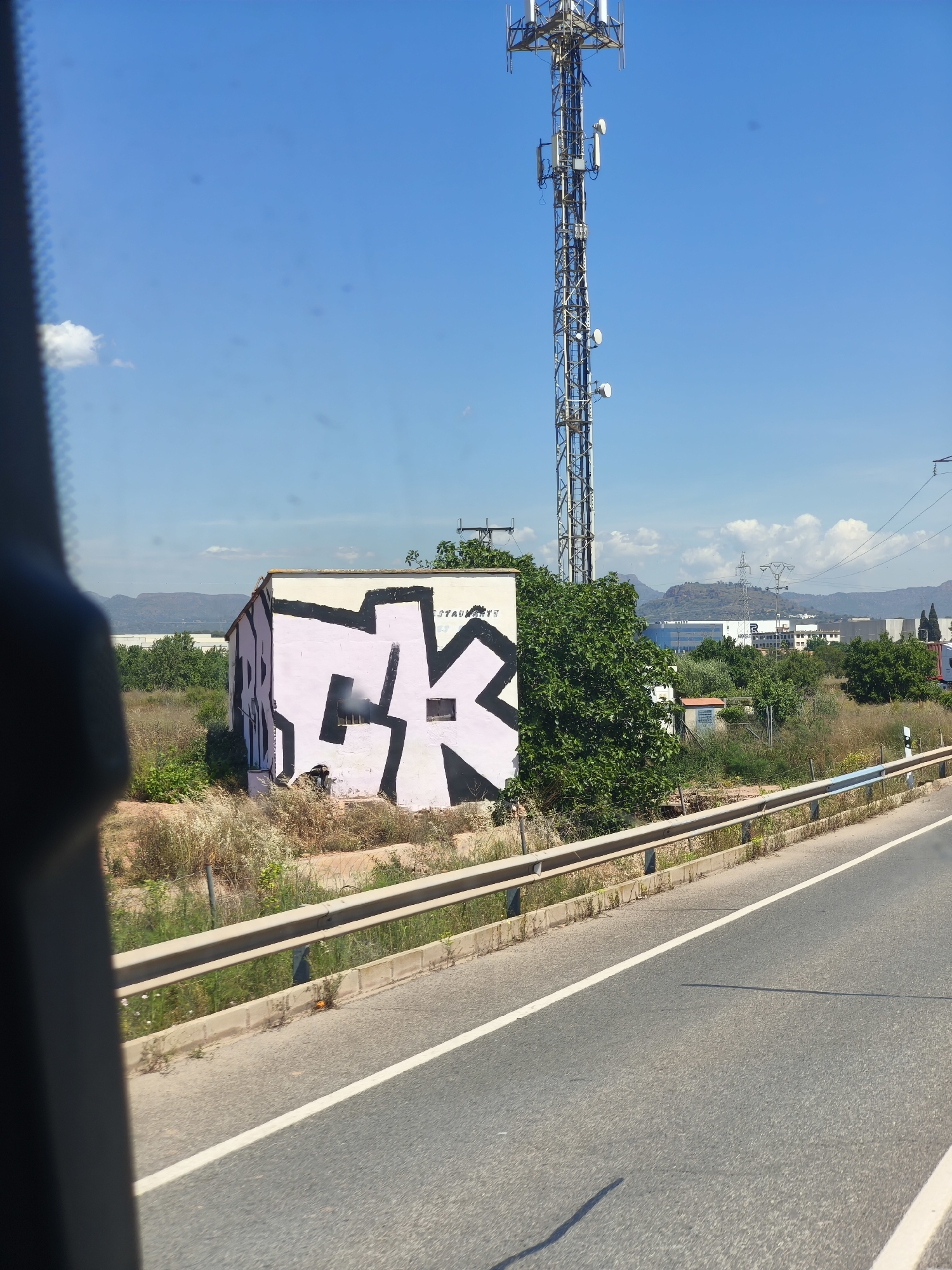 The image shows a roadside scene in Museros, Spain. A cell tower rises high in the background against a clear blue sky. In the mid-ground, a small, rectangular building with graffiti (predominantly a large, stylized, pink and black "K") is visible, flanked by vegetation. A metal guardrail runs parallel to the road. The road itself occupies the foreground, marked with a white lane divider. A dark object, possibly a window frame, is visible on the left edge of the frame. No people are visible.