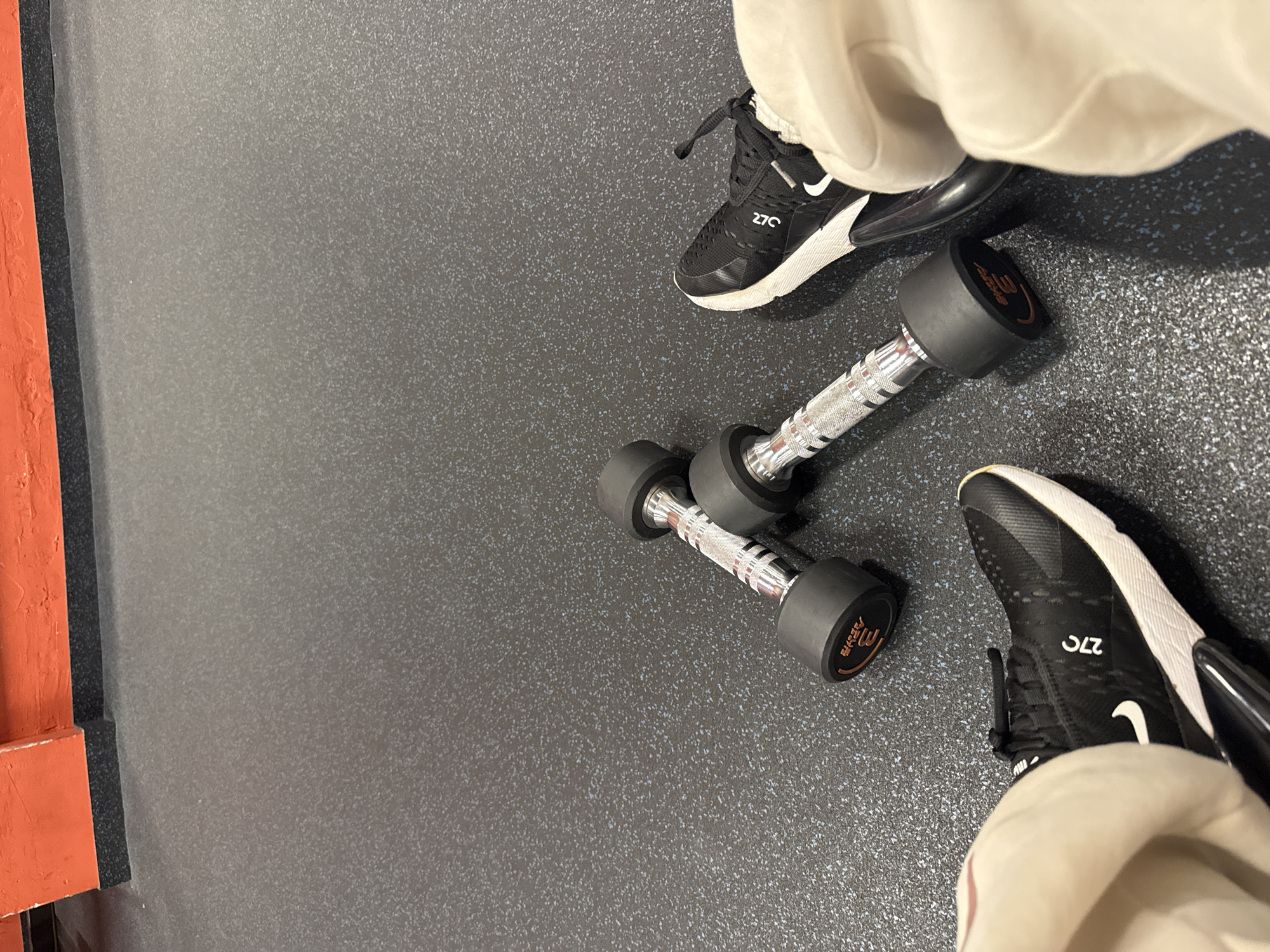 The image depicts an indoor scene within a fitness facility in Sofia, Bulgaria, captured from a first-person, downward-looking perspective. In the immediate foreground, a pair of feet wearing black Nike Air Max 270 sneakers with white soles are visible, positioned at the bottom left and right corners of the frame. The shoes are partially obscured by light-colored pants. Centrally positioned between the feet are two identical fixed-weight dumbbells, each featuring black rubberized hexagonal ends and a chrome, knurled handle. The end cap of one dumbbell clearly displays "3 KG" and the brand "SKSPORTS". The floor beneath the subjects is a dark grey rubber matting, speckled with white and light blue particles, commonly used in gyms. In the upper portion of the frame, an orange-painted wall with a distinct black base trim is visible, suggesting the perimeter of the room. The lighting is uniform, indicative of artificial indoor illumination.