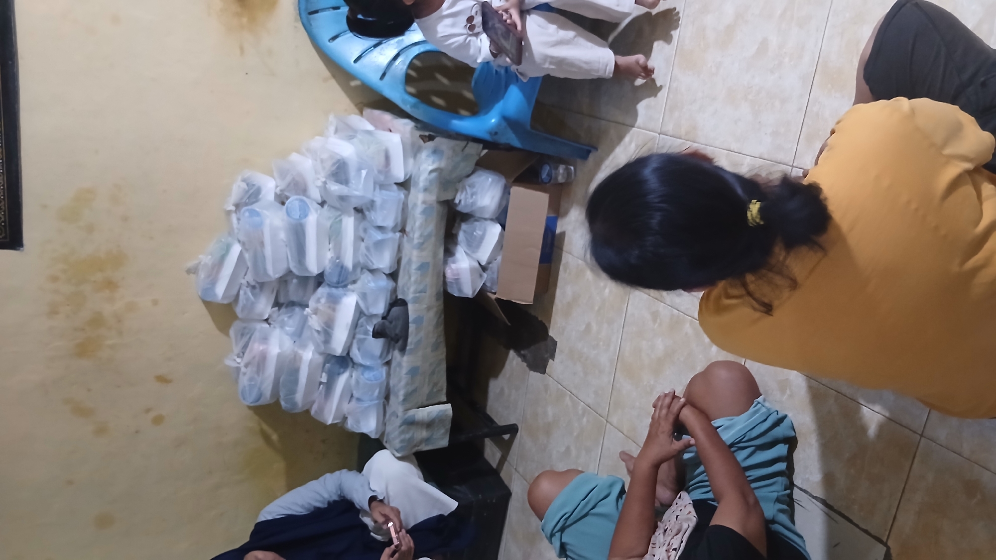 In the city of Klambir Lima Kebun, Indonesia, a group of individuals is gathered in what appears to be a home setting. The scene is dominated by a large pile of food containers, wrapped in plastic, stacked on a table covered with a patterned cloth. These containers suggest a meal distribution or preparation event.

Several people are visible in the image. To the left, a person wearing a dark blue headscarf and a light-colored top is seated. In the foreground, two individuals are seated on the floor, their backs to the camera. One is wearing a light blue garment, and the other is in a bright yellow shirt. To the right, a person in white clothing is seated on a blue plastic chair, holding a tablet or similar device. Their legs are visible, indicating they are likely a child.

The environment appears to be a simple interior, with a beige wall as the backdrop, marked by some water stains. The floor is tiled in a beige, square pattern. A cardboard box is placed on the floor near the table, and a few water bottles are also visible. The overall atmosphere suggests a communal gathering, possibly for a charitable purpose or a family event.