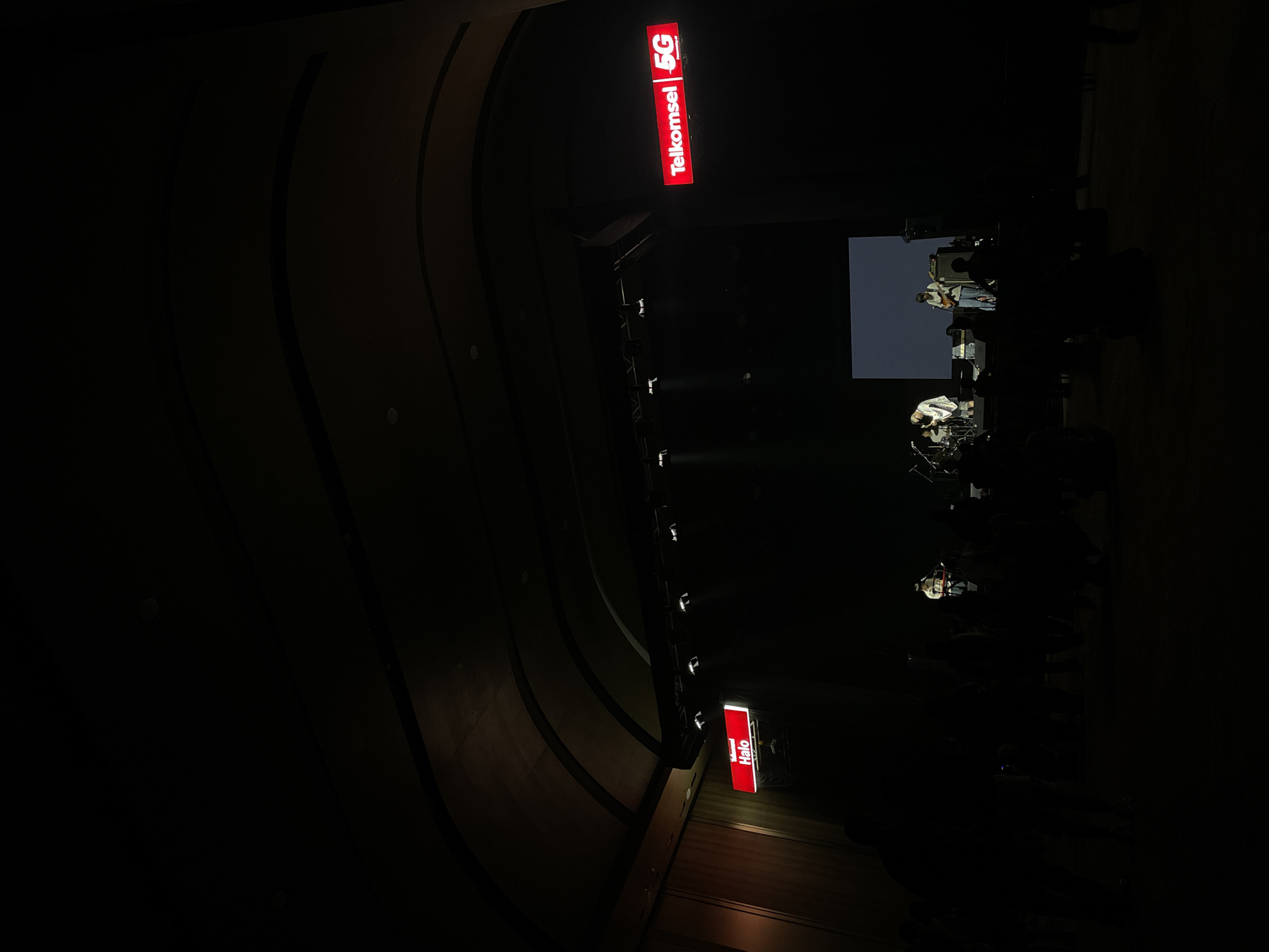 The image shows a dimly lit indoor space in Salembaran, Indonesia. A stage with musicians performing is visible under spotlights, with a screen behind them. Two large, illuminated signs for "Telkomsel 5G" and "Telkomsel Halo" are positioned above the stage. The walls have a curved, textured design. The overall environment is dark, with only the stage and signs brightly illuminated.
