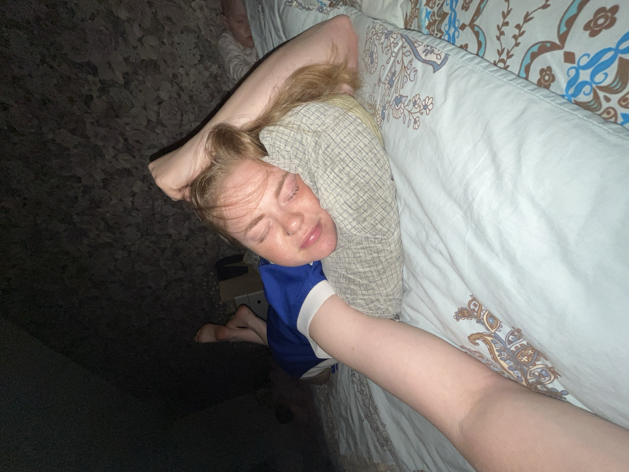 A fair-skinned person is lying on their side in a bed with patterned bedding. The person's face is visible; their eyes are closed and they appear to be sleeping. Their blonde hair is pulled back and partially covered by a patterned shirt or blanket. A portion of a blue shirt with a white sleeve is visible on their torso. Their arm is extended along the length of the bed. The background shows a dark, carpeted floor. There is no information to suggest that the location is the city of Chaikovsky, Russia.