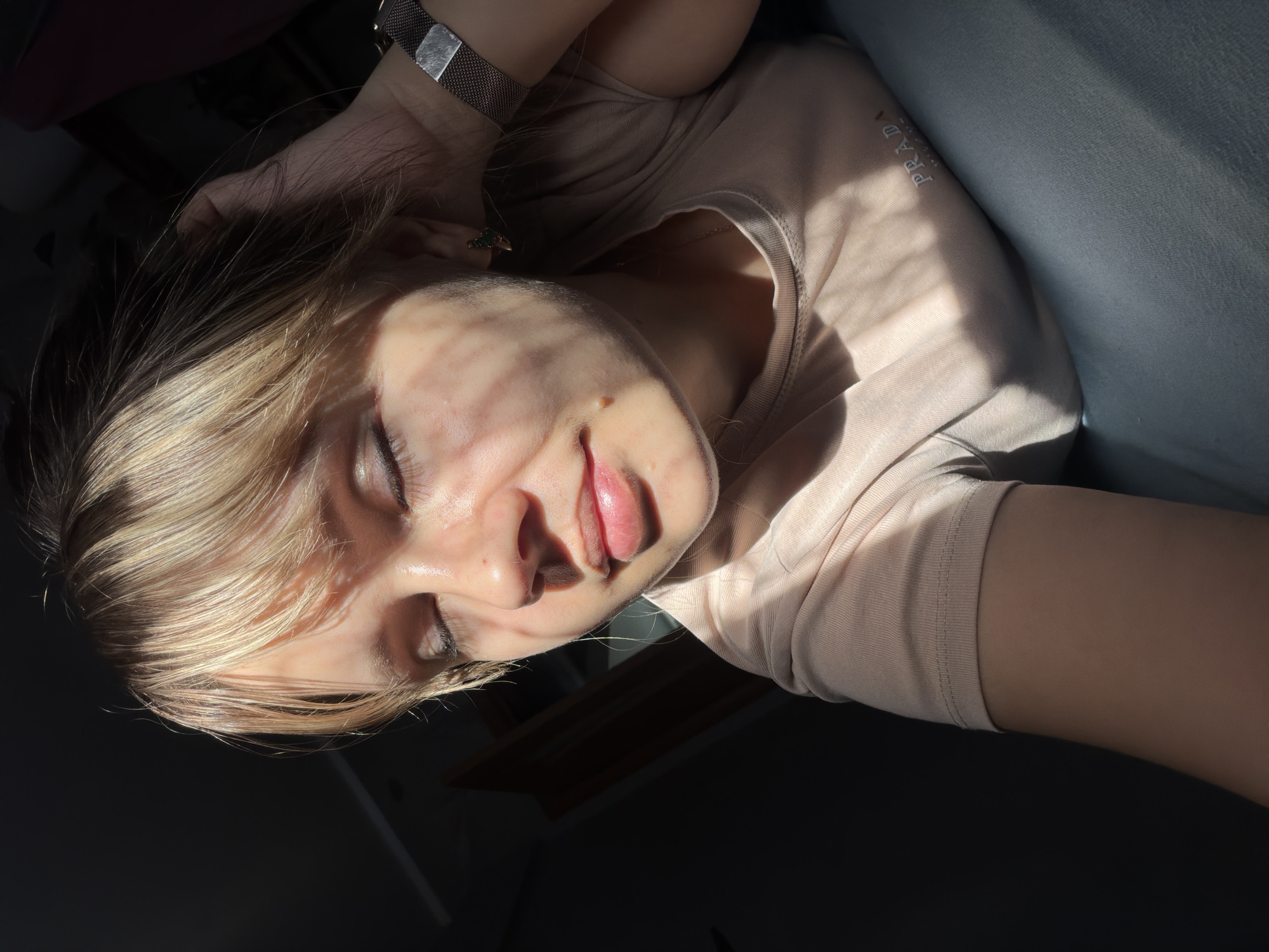 An intimate, close-up shot features a person lying down, illuminated by strong, direct sunlight. The person has blonde or light brown hair, with several strands falling across their forehead and partially obscuring their face. Their eyes are closed, and their lips are slightly parted, conveying a sense of relaxation or contemplation.
Horizontal shadows, likely cast by window blinds or slats, create a distinct striped pattern across the person's face, chest, and arm. They are wearing a light beige or pale pink short-sleeved t-shirt, on which the word "PRADA" is visibly embroidered in white or light thread on the left chest area. A metallic mesh watch is worn on their left wrist, and a small ring, possibly with a green stone, is on their left hand, which is positioned behind their head.
The background is predominantly dark, suggesting a shadowed indoor space, with a lighter, possibly fabric-covered surface visible to the right. The strong highlights on the skin and hair indicate a bright light source, likely a window, suggesting the time of day is either morning or late afternoon. The overall mood is peaceful and serene.