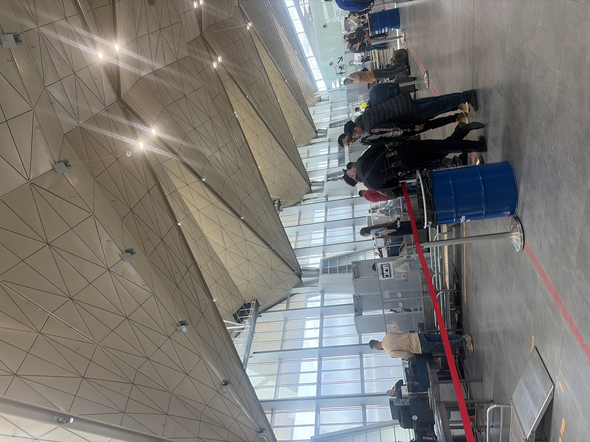 The image, rotated 90 degrees clockwise, captures an indoor scene at what appears to be a modern airport or large transport hub in Saint Petersburg, Russia, likely during daytime given the bright natural light. The most striking architectural feature is the expansive, geometric ceiling composed of numerous triangular panels, some illuminated by embedded lights creating distinct starburst patterns.
A queue of people, mostly adults dressed in casual attire including jackets and jeans, stretches across the right side of the frame, moving towards a security screening area. Individuals are seen waiting patiently, with backpacks and other luggage visible. In the foreground, a person in a beige top and jeans stands near a red retractable barrier, facing towards the screening equipment. Further along the queue, security checkpoints with X-ray machines and metal detectors are discernible.
The floor is a plain grey, marked with red and yellow lines, guiding the flow of the queue. Blue metal barrels, possibly used as waste bins or temporary bollards, are interspersed along the queuing area. Silver stanchions with red retractable belts form the queue barriers. Large, multi-pane windows span the upper portion of the wall on the right, allowing ample sunlight into the facility. A small sign with an arrow pointing right and a stylized person icon is visible above the queue, likely indicating direction or a facility. Text on other signs and the blue barrels is too indistinct to read. The overall atmosphere appears orderly, consistent with passengers undergoing security procedures.