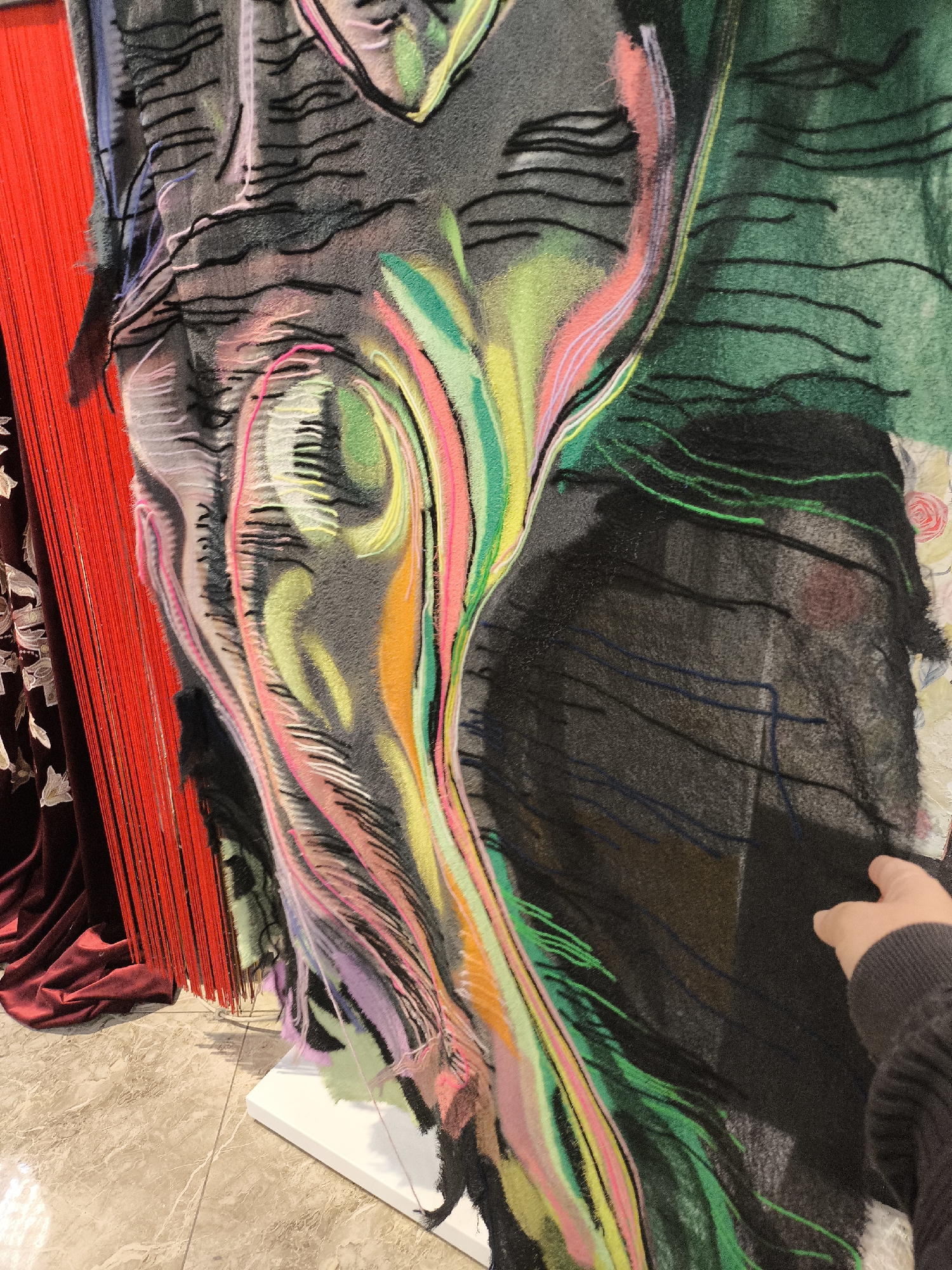 The image displays a vibrant, abstract textile artwork exhibited indoors, likely within a gallery or showroom in Khimki, Russia. The central piece is a large textile, predominantly dark grey or black, adorned with dynamic, undulating shapes and lines. These colorful patterns are created using various yarns or threads in shades of pink, green, yellow, orange, purple, and blue, providing a vivid contrast against the darker base fabric. Black lines, resembling stitches or drawn marks, are also integrated into the design. The artwork's texture appears soft and tactile, suggesting techniques like tufting or embroidery.
To the left, a dense arrangement of vertical red fabric strands, resembling a curtain or tassels, provides a strong visual accent. Below these, dark red or maroon draped fabric with a subtle patterned texture is visible. The textile artwork rests on a white stand, positioned on a polished, light-colored tiled floor with a marble-like pattern.
A human hand, wearing a dark sleeve, extends into the frame from the right, appearing to gently touch or interact with the textile art. This suggests active engagement or examination of the artwork by a viewer. The scene is evenly lit by indoor lighting. There is no visible text in the image. The overall impression is one of a contemporary and expressive textile art display.