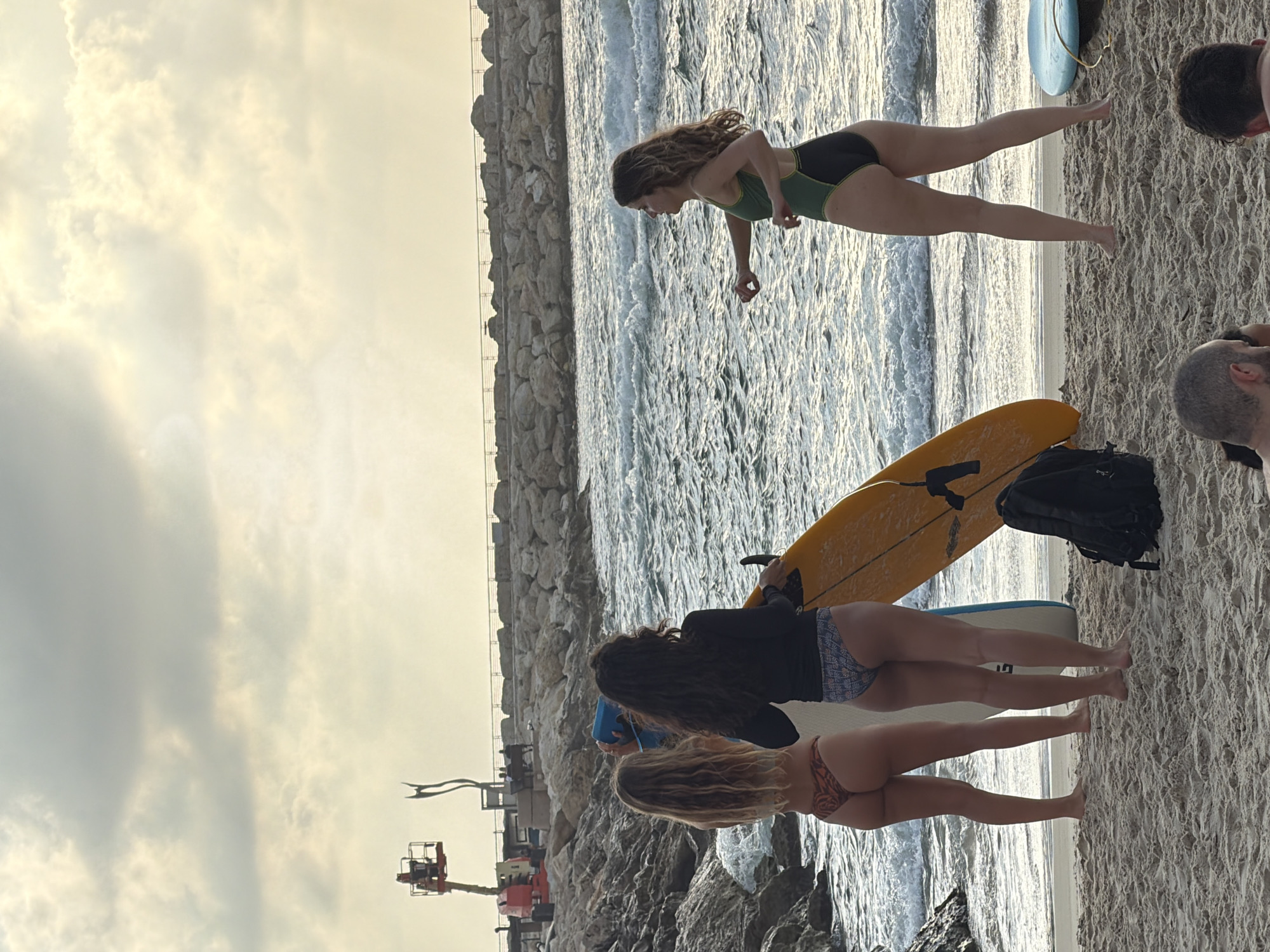 Three young women, two in bikinis and one in a swimsuit, stand on a sandy beach next to surfboards. The woman on the far left wears a patterned bikini. The middle woman wears a dark long-sleeve top and patterned bikini bottoms. The woman on the right wears a green and black swimsuit. They are near the water's edge, with waves breaking on the shore. In the background, a rocky breakwater extends into the sea, with a construction crane and a metal sculpture visible on the shoreline behind it. The sky is overcast with clouds. This scene appears to be at Gordon Beach in Tel-Aviv, Israel, given the distinctive breakwater and the presence of surfboards.