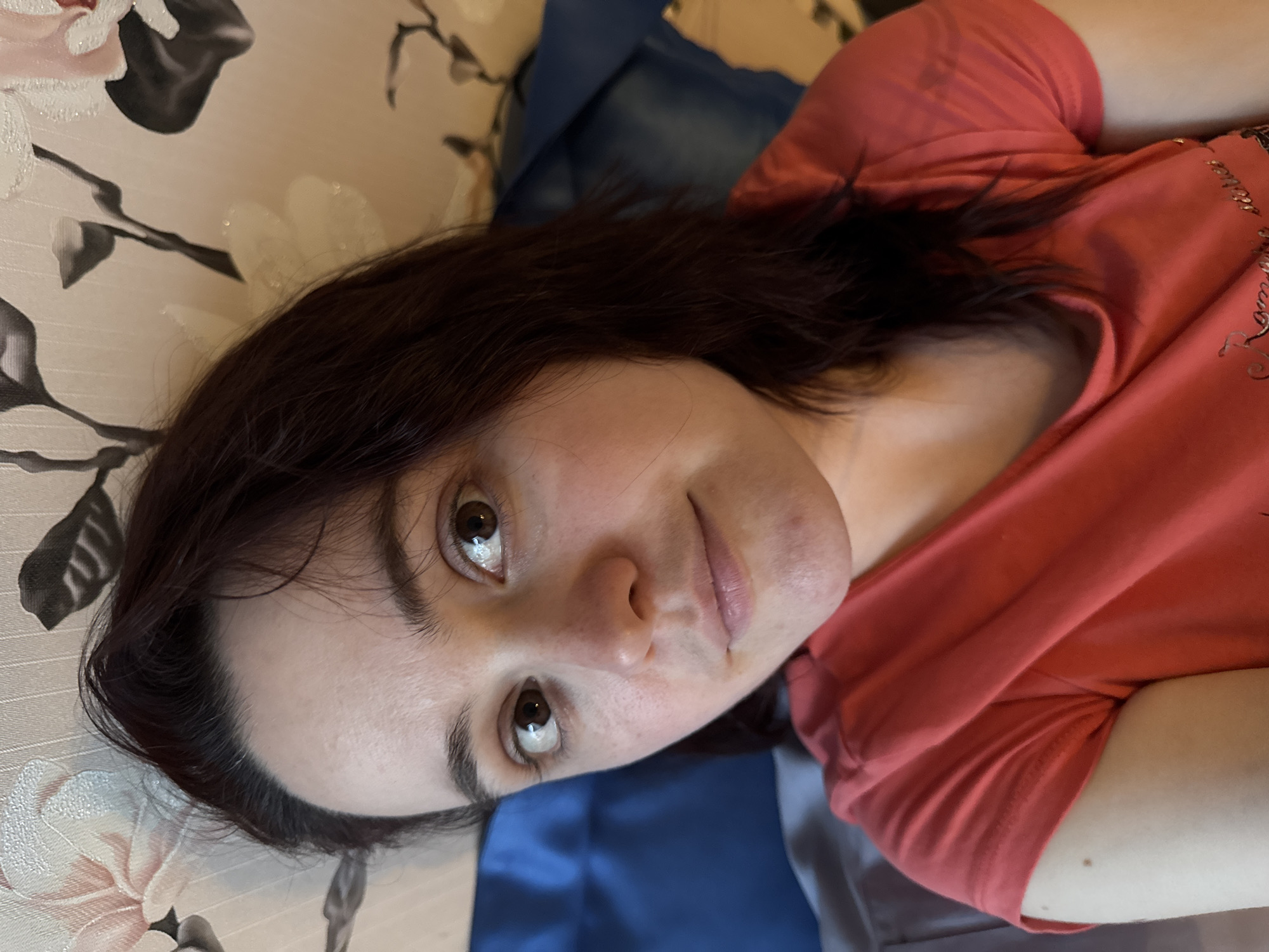 A young woman with dark hair and light skin is lying on a surface with a floral pattern, likely a bed or couch. She is wearing a red t-shirt. The background includes a blue object or surface. The image does not contain enough information to determine the location as Sochi, Russia.