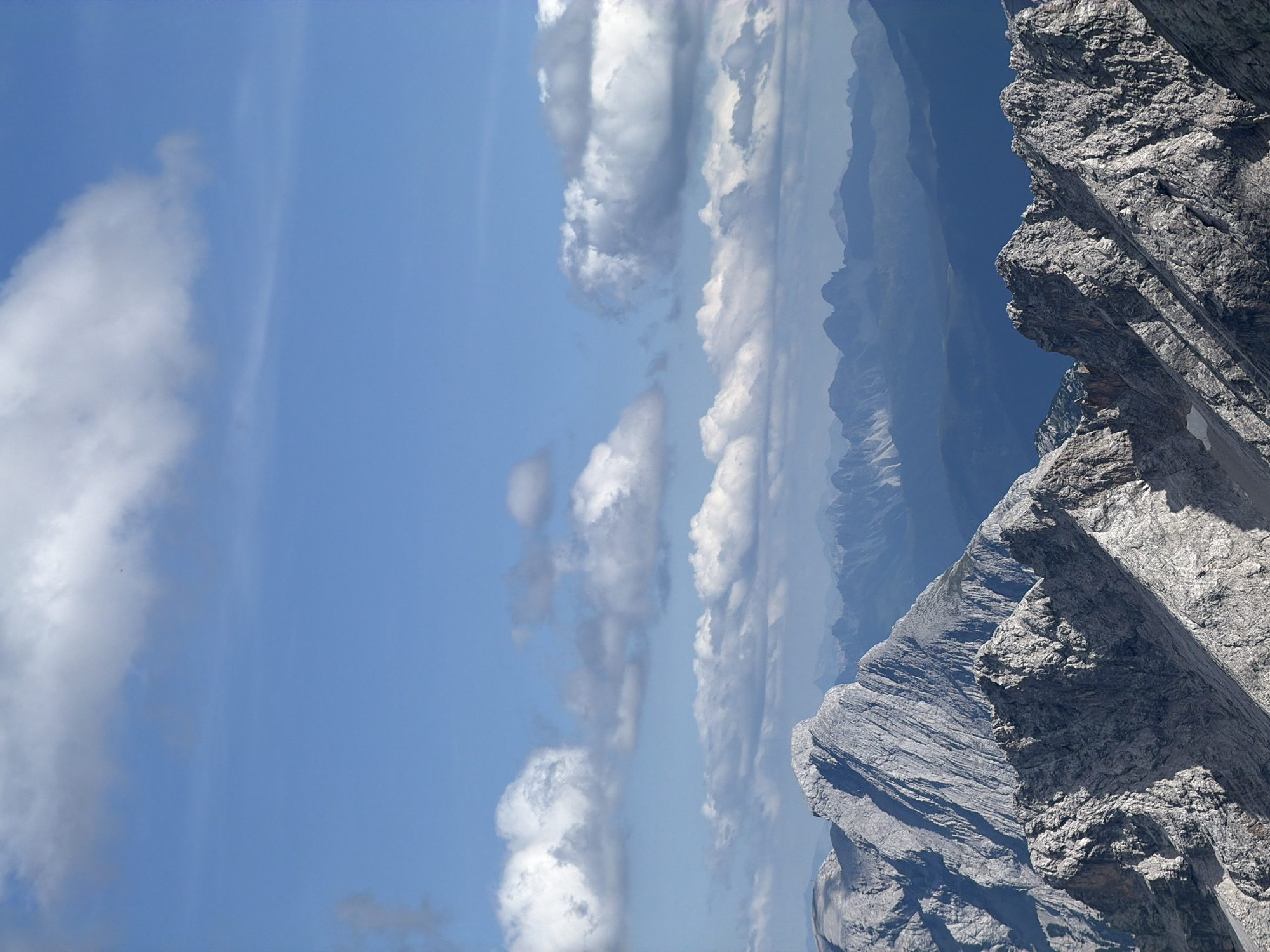 The image depicts a mountainous landscape under a partly cloudy sky. The foreground is dominated by rugged, rocky mountain ridges with sharp, jagged peaks. The rock faces are textured and show signs of erosion, with shadows cast across their surfaces. In the middle ground, more mountain ranges are visible, appearing softer and more distant with a bluish hue, indicating atmospheric perspective. The background consists of a clear blue sky with scattered white cumulus clouds. There are no people, animals, or man-made objects visible in the image. The scene is a natural mountain environment.
This view is consistent with vistas from the Zugspitze, Germany's highest peak, which is located near Garmisch-Partenkirchen.