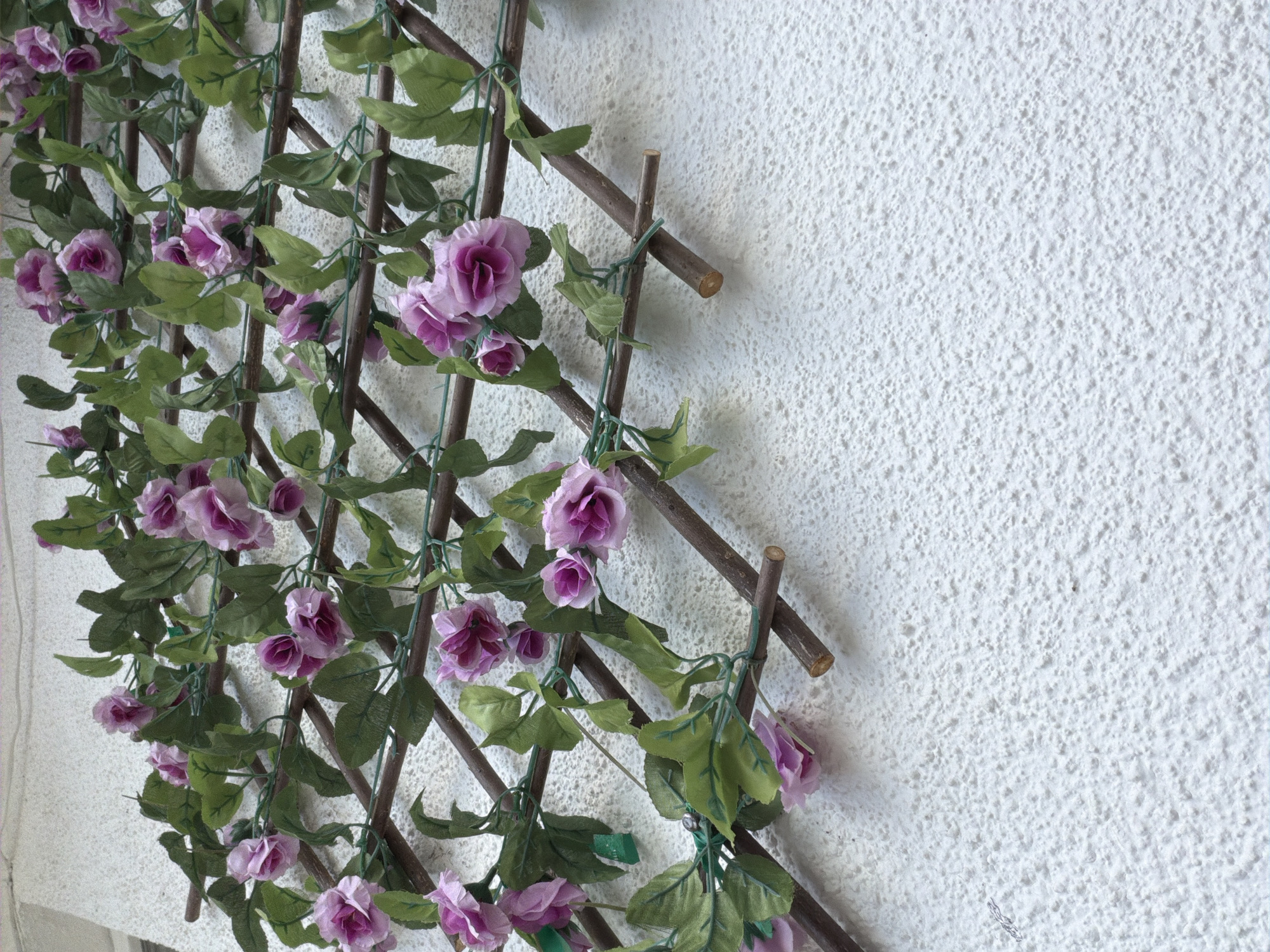 This media file is a close-up, eye-level shot of a decorative trellis covered in artificial purple flowers and green leaves, leaning against a textured, white wall. The trellis is made of thin, brown wooden sticks forming a diamond pattern, secured with green ties. The flowers are clustered along the trellis, some fully open and others in bud, with a gradient of pink and purple petals. The green leaves are also artificial and spread out, partially obscuring the trellis structure. The white wall behind the trellis has a rough, stucco-like texture with many small, raised bumps.
The scene is static, with no discernible activity taking place. The lighting suggests it is daytime, possibly outdoors due to the texture of the wall which could be an exterior surface. The image lacks any people or clear indicators of specific time of day or weather conditions. The location context of Tbilisi, Georgia, does not provide specific visual cues within the image itself. There is no visible text in the image. The overall impression is of a decorative element in an outdoor or semi-outdoor setting, possibly a garden or balcony.