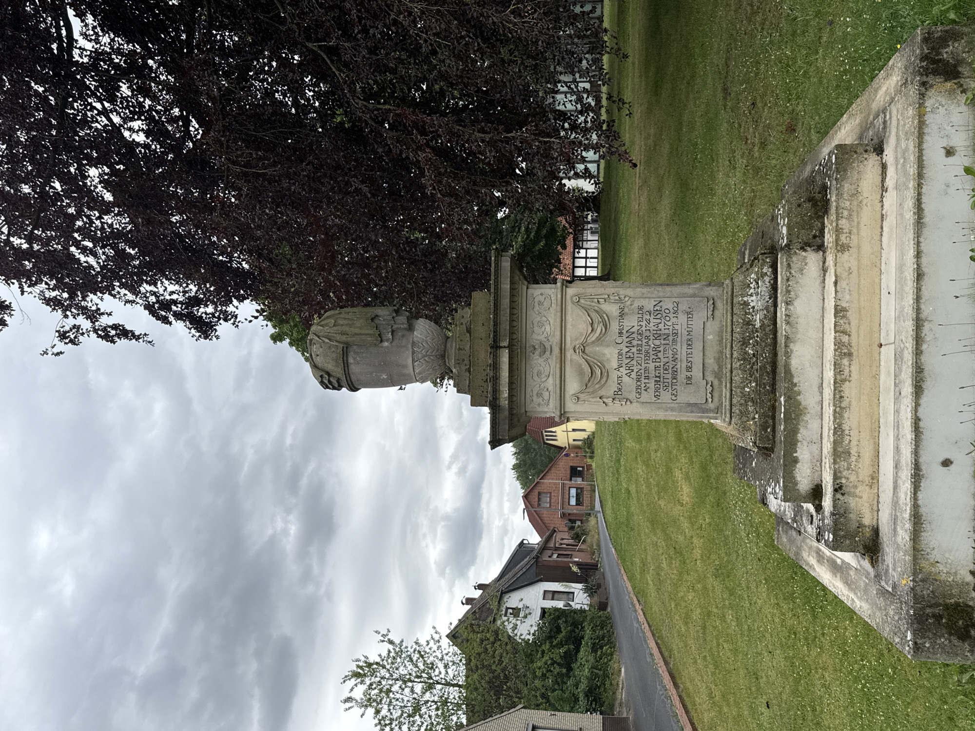 This is a vertical, eye-level, medium shot of a monument with text inscribed on it, set against a backdrop of a cloudy sky, a large tree with dark foliage, and a grassy area with some houses and trees in the distance. The monument appears to be made of stone, with a carved urn-like feature at the top. The inscription on the monument is in German and appears to be a dedication or memorial. The text includes names and dates, suggesting it is a gravestone or memorial plaque. The overall mood of the image is somber, possibly due to the overcast weather and the nature of the monument. The location appears to be a public space, likely a cemetery or a park, given the presence of the monument and the surrounding greenery. The houses in the background indicate a residential or village setting. The image was likely taken during the daytime, given the visibility of the scene.