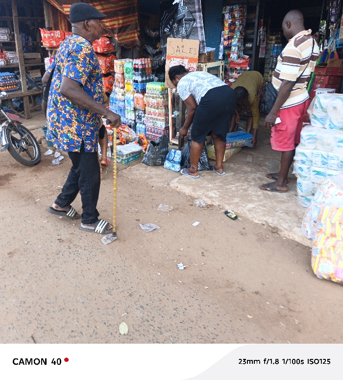 A street scene features an elderly man in a patterned shirt and black cap, using a cane while walking. A small motorcycle is parked behind him. In the background, several people are shopping at a roadside stall displaying various beverages and household items. One person is reaching into a display of goods, and another stands nearby. A "SALES" sign is visible. Litter scatters the ground in the foreground. The scene is captured in daylight, with a busy, everyday atmosphere.