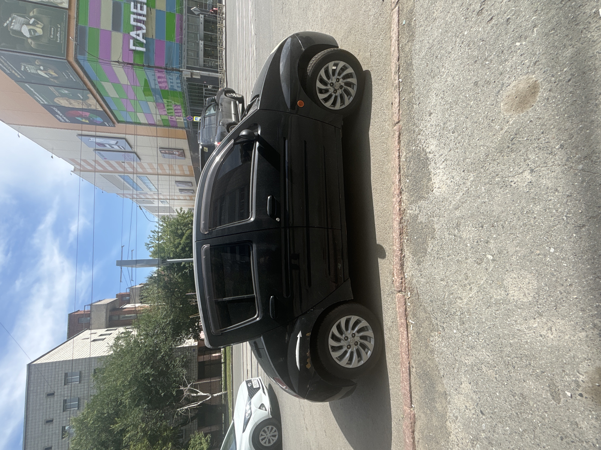 The image shows a black car parked on the side of a street in Novosibirsk, Russia. The car is a small hatchback with tinted windows and silver alloy wheels. It is parked on an asphalt surface, with a red curb visible in the foreground. In the background, there are buildings, trees, and other cars. One of the buildings appears to be a shopping mall with colorful panels and large advertisements. The sky is blue with some clouds. The scene suggests a typical urban environment on a sunny day.